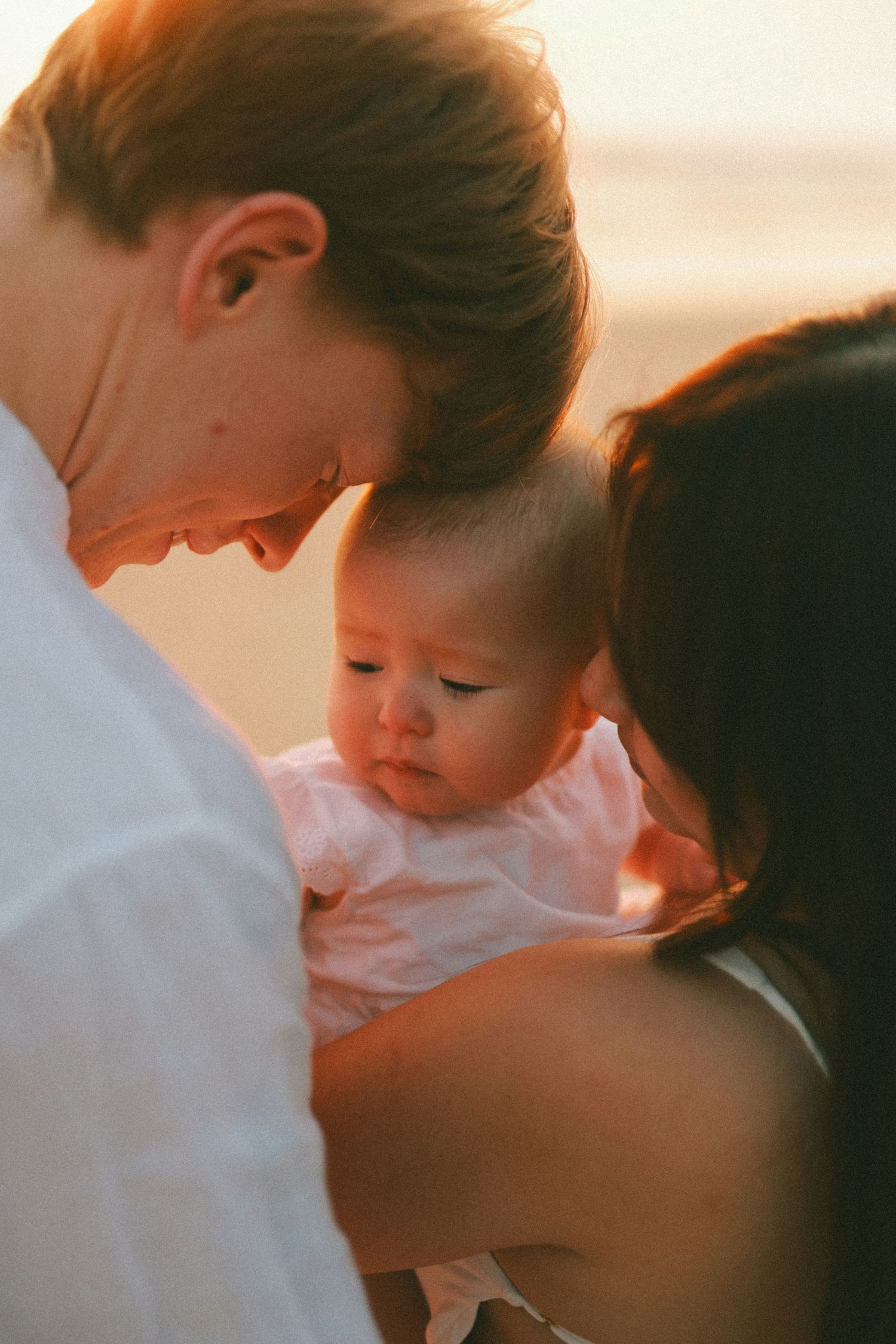 Parents leaning heads together over their baby girl in pink dress during golden sunset outdoors.