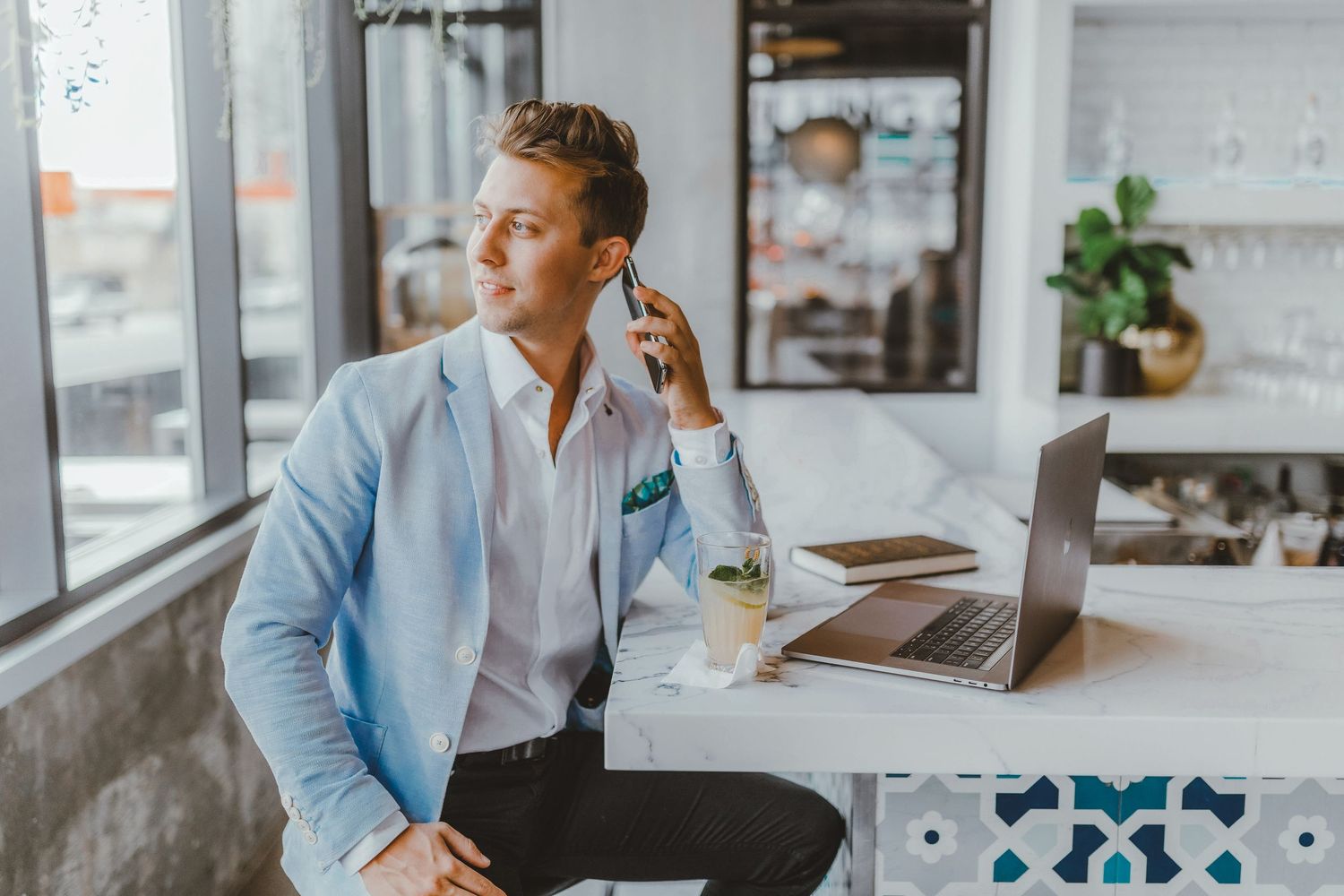Young businessman in light blue blazer talking on phone while working on laptop in cafe.