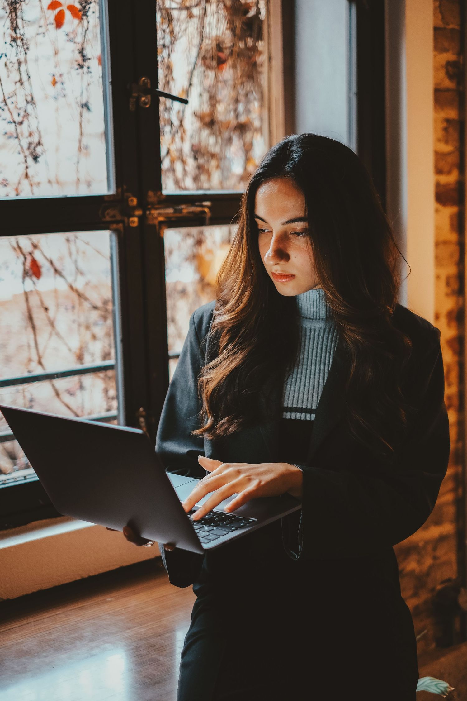 Young professional woman in black blazer using laptop while standing near autumn window.