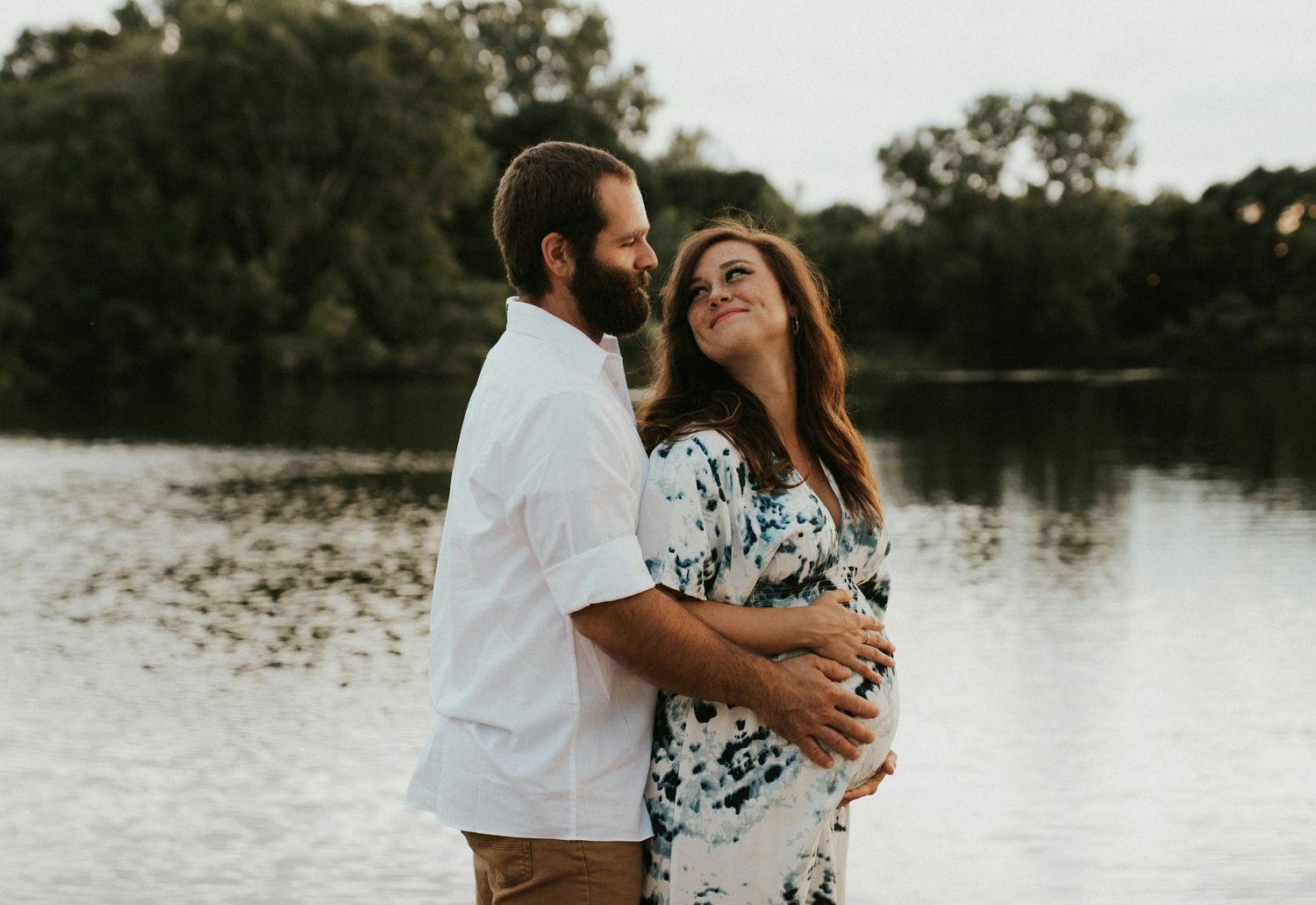Smiling pregnant couple embracing near a lake, woman in tie-dye dress, man in white shirt at dusk.