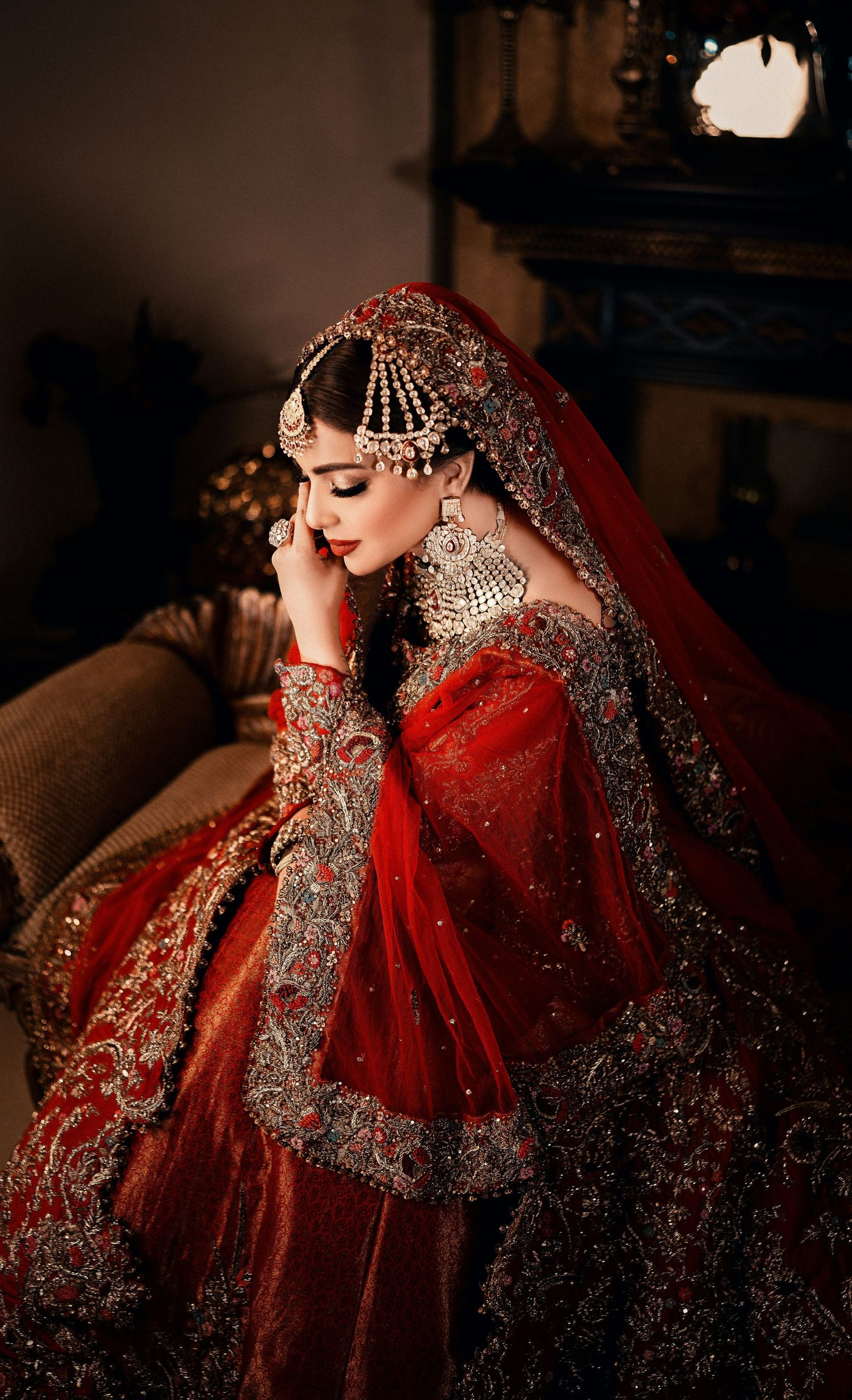 Stunning bride in heavily embroidered red Pakistani bridal lehenga seated in dramatic lighting.