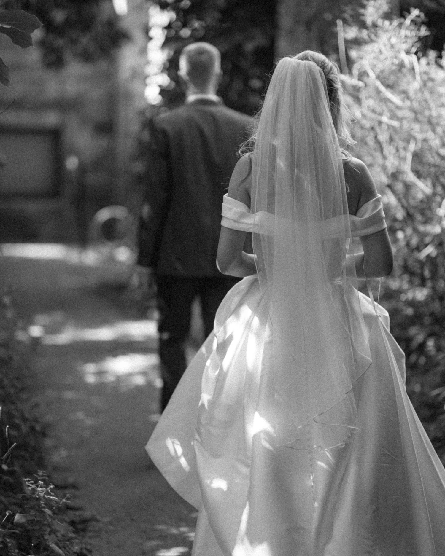 Black and white photo of bride in veil and gown walking with groom along garden path.