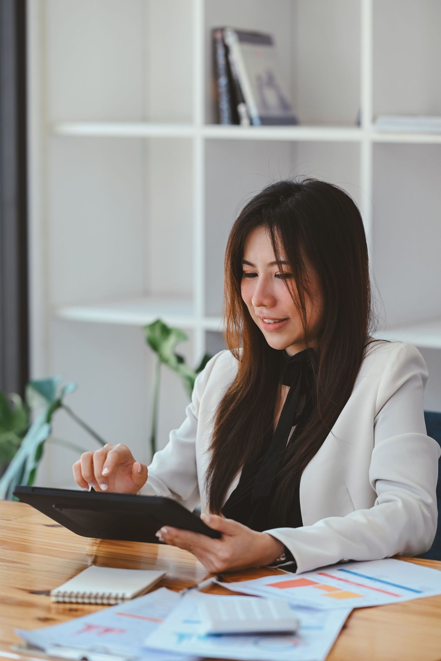 Asian businesswoman in white blazer using digital tablet while reviewing charts at office desk.