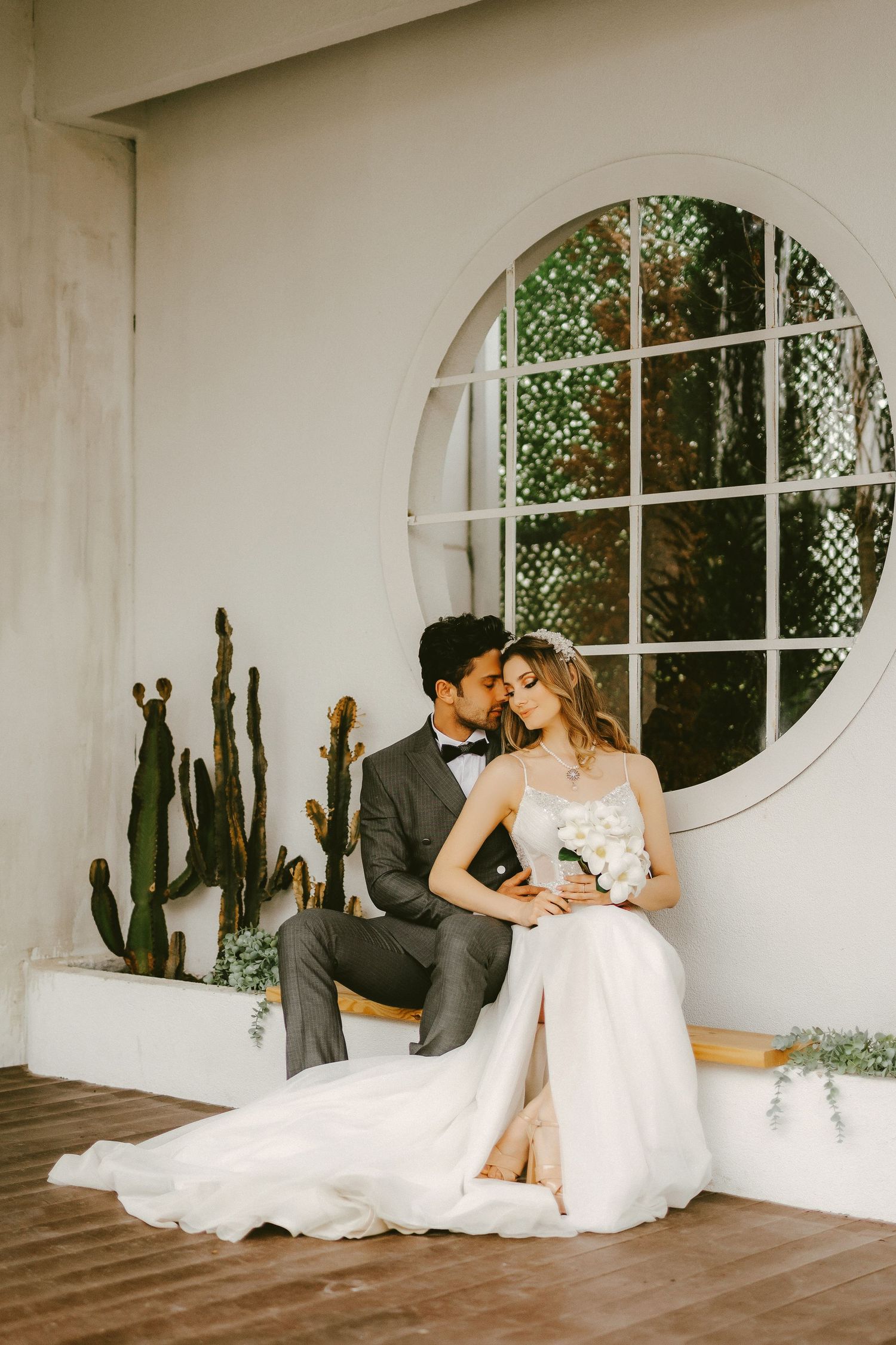 Bride and groom share romantic moment seated before round window with cacti backdrop on wedding day.