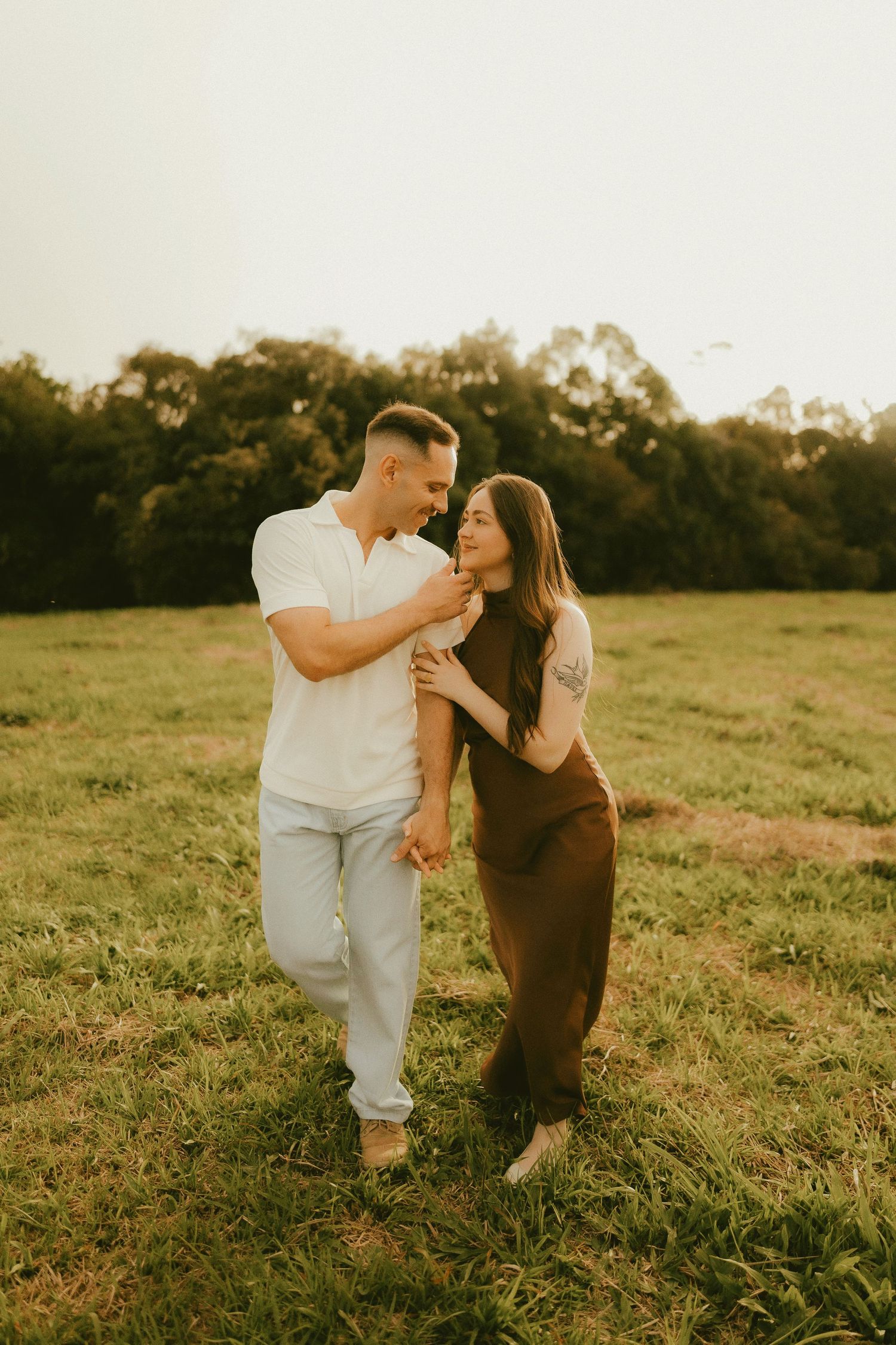 Couple sharing a romantic moment in a sunlit green field, man in white shirt and woman in brown dress holding hands.