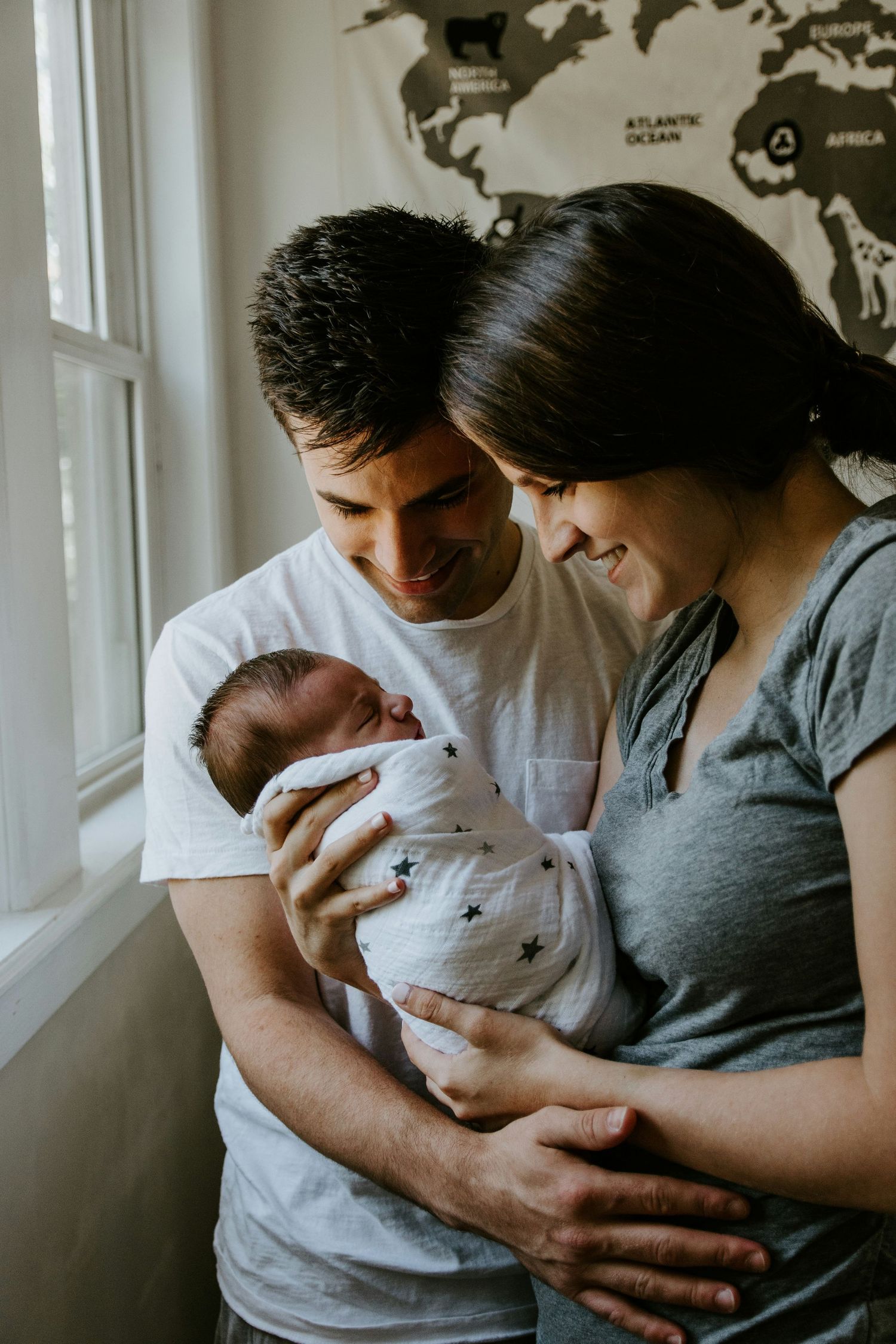 New parents smile warmly while holding their swaddled newborn baby together near a bright window.