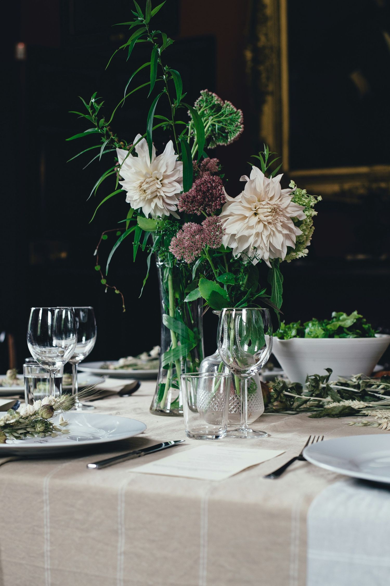 Elegant wedding reception table set with white dahlias, greenery, crystal glasses, and linen tablecloth.