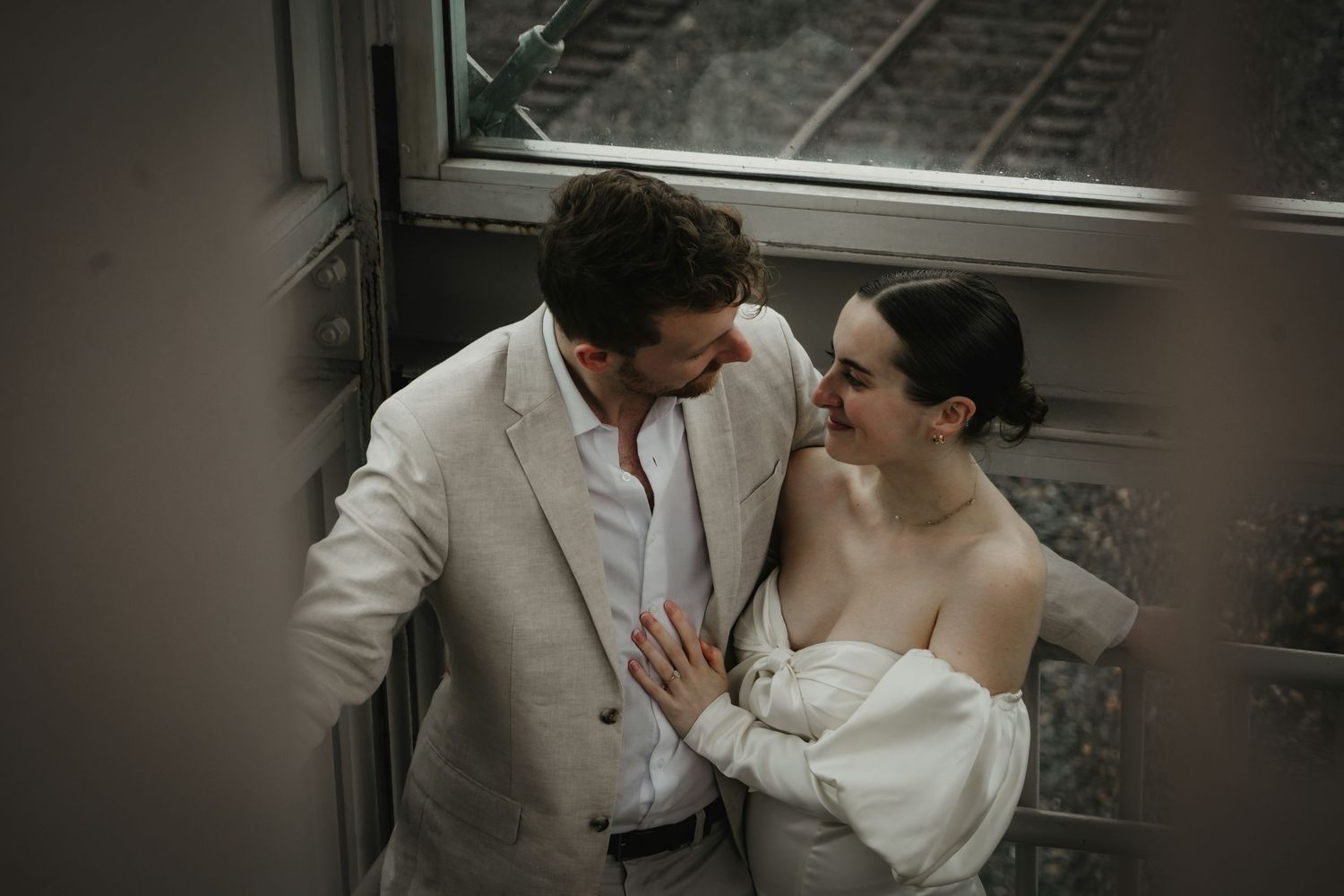 A bride and groom share an intimate moment leaning against a window with train tracks visible in the background.
