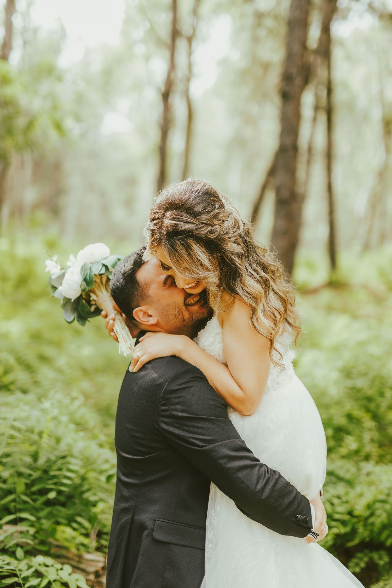 Bride and groom sharing a romantic kiss in a lush green forest, bride holding white bouquet in wedding attire.