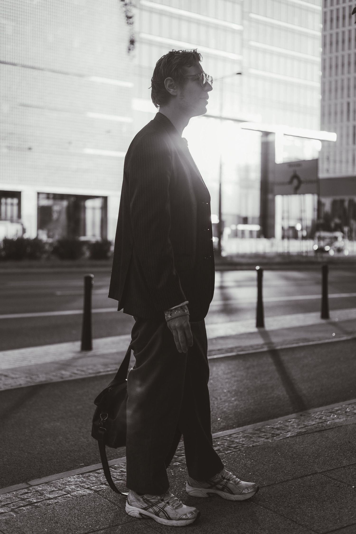 Black and white photo of person in dark outfit standing on urban sidewalk.