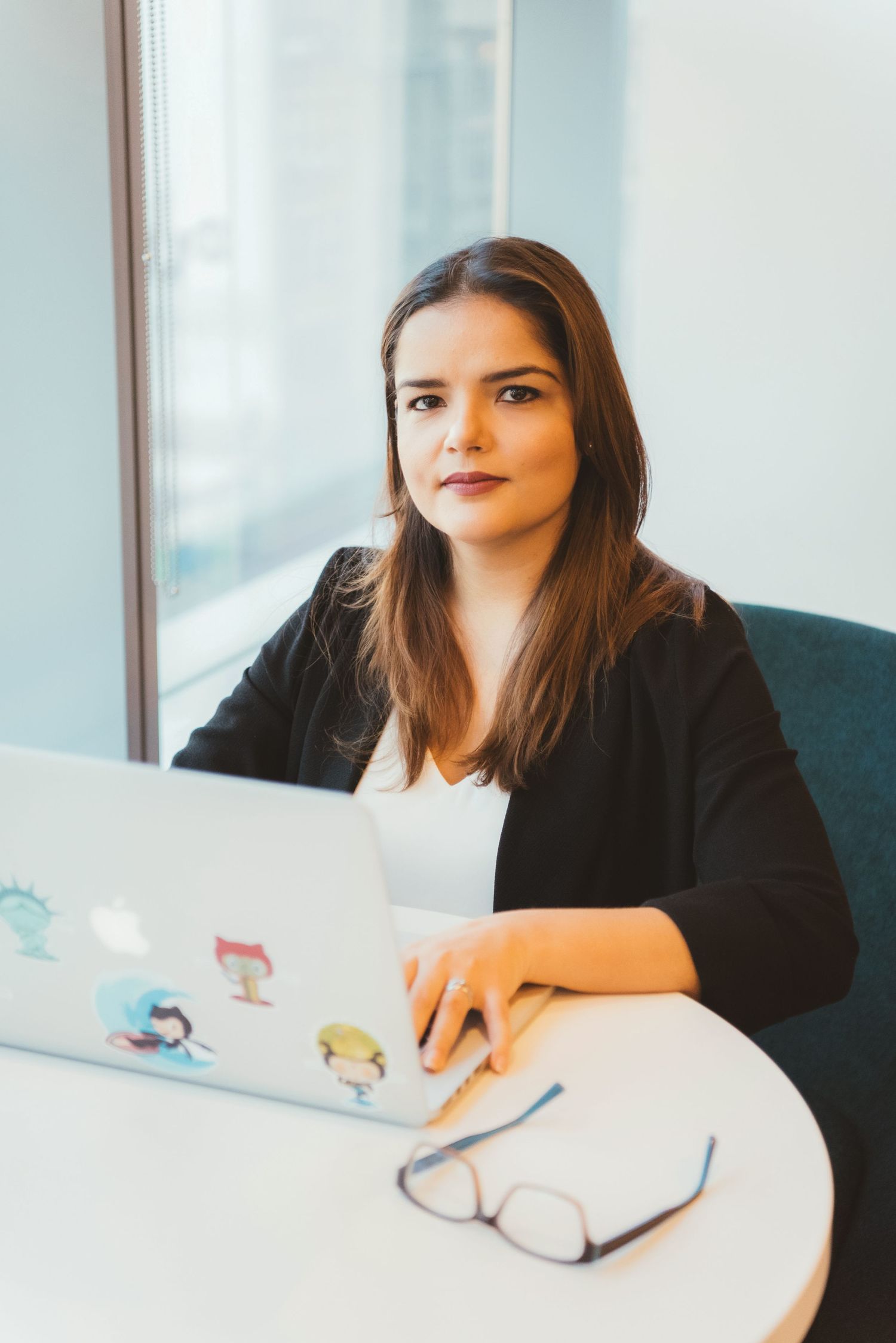 Professional brunette woman in black blazer working on sticker-covered laptop at white office desk.