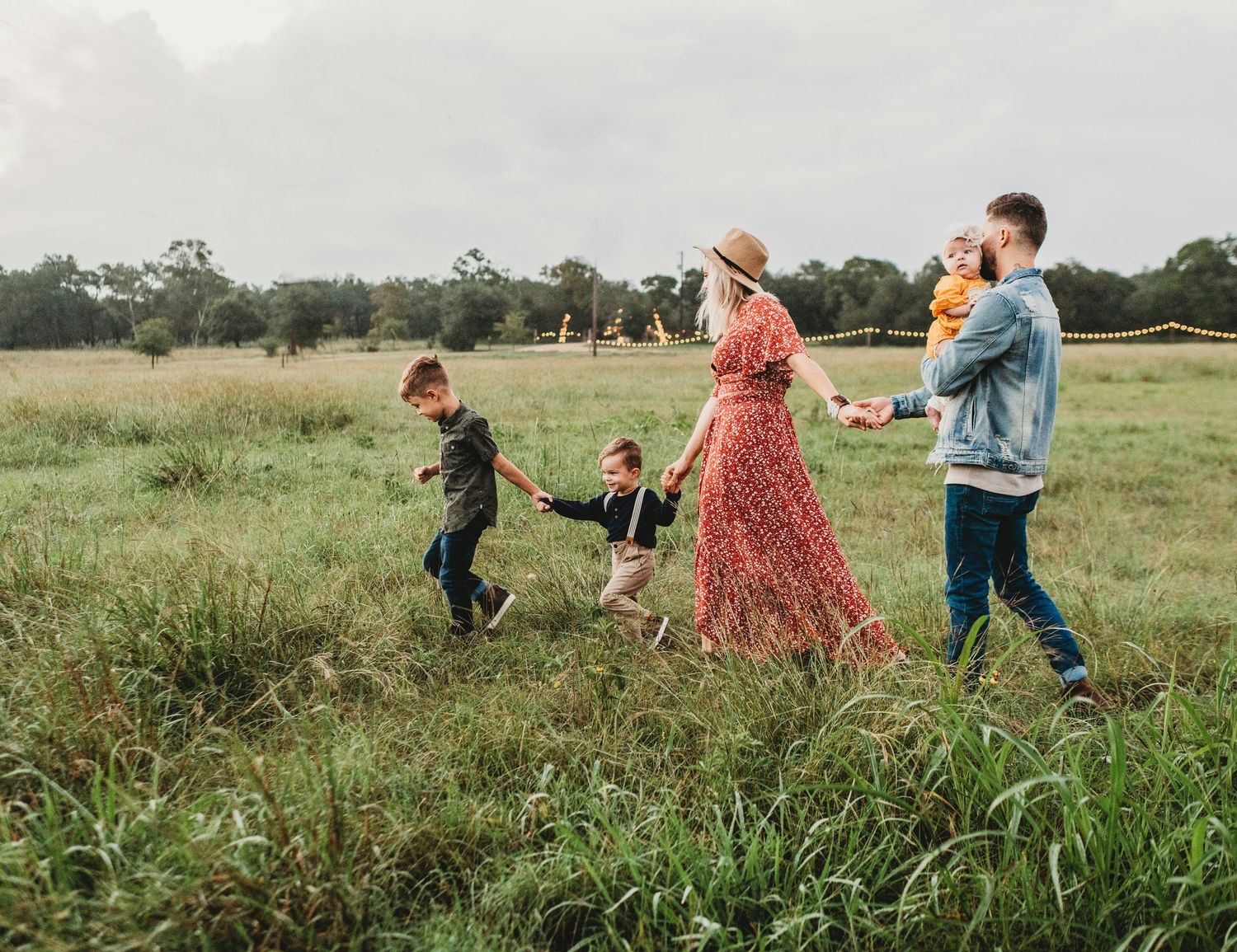Family of five walking through a lush green meadow, parents holding children's hands on a cloudy day.