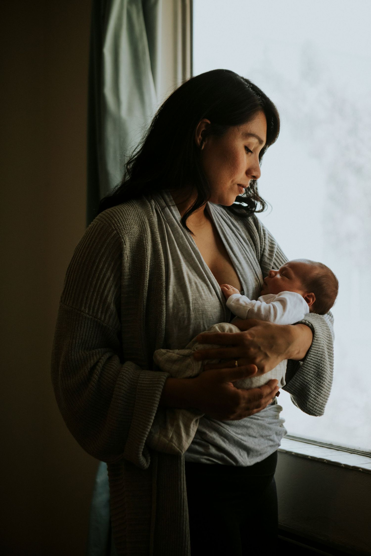 Mother standing by window in grey cardigan tenderly cradling her newborn baby in soft natural light.