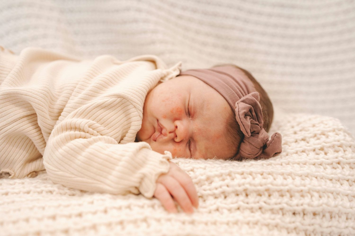 Sleeping newborn baby girl in cream knit outfit and floral headband on white blanket.