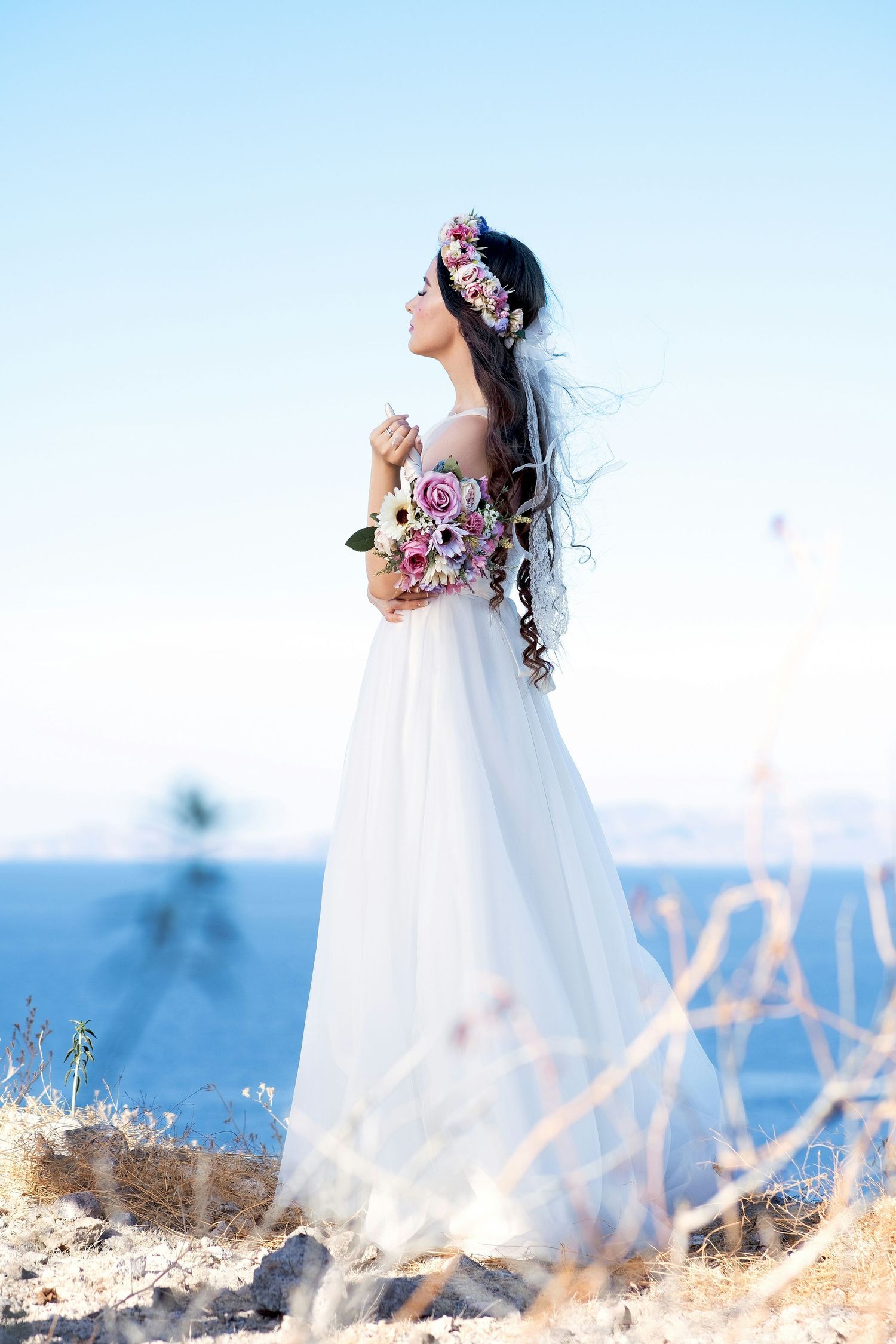 Bride in white flowing gown with floral crown holds bouquet by the sea.