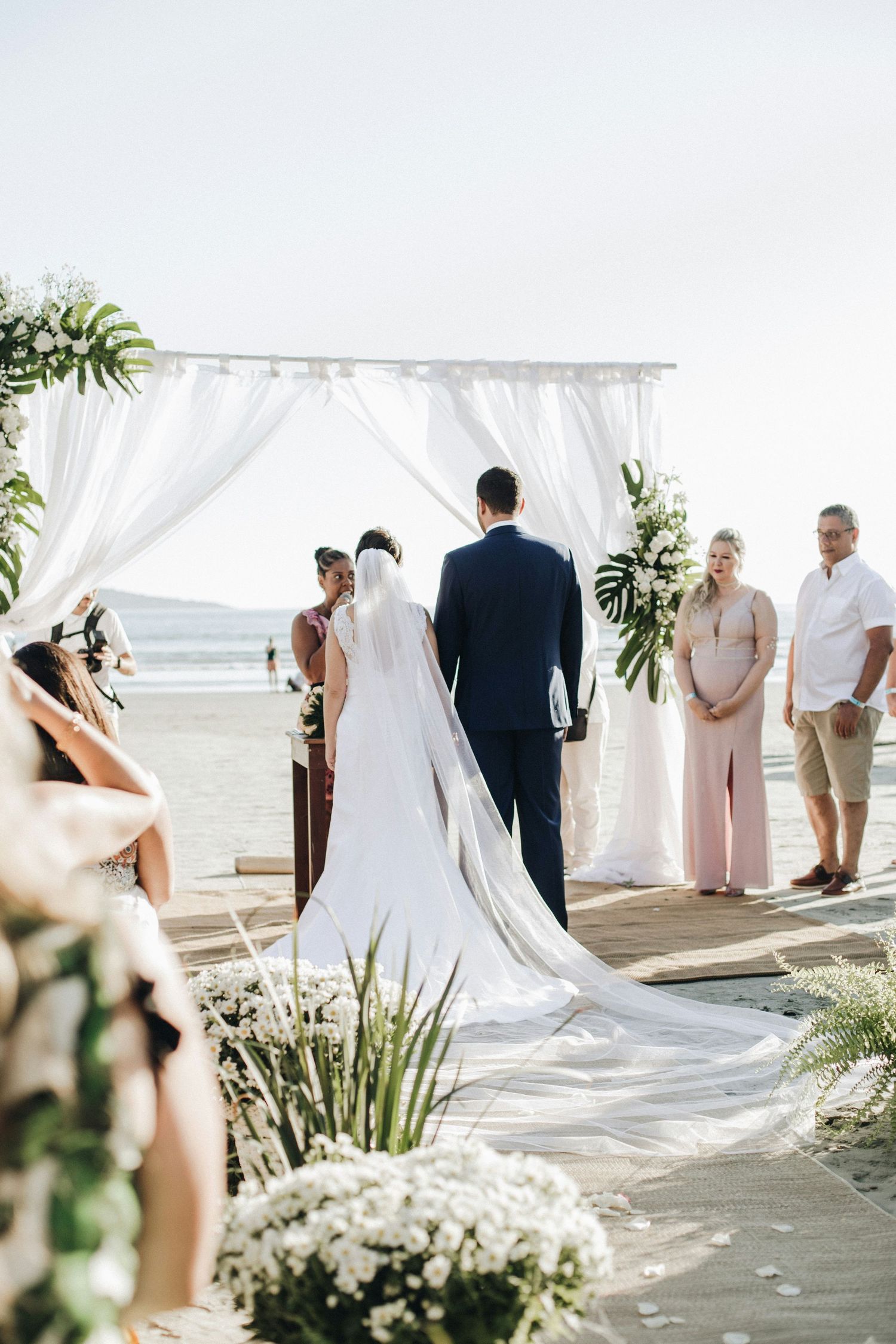 Bride and groom exchanging vows at elegant beach ceremony under white draped arch with tropical florals.