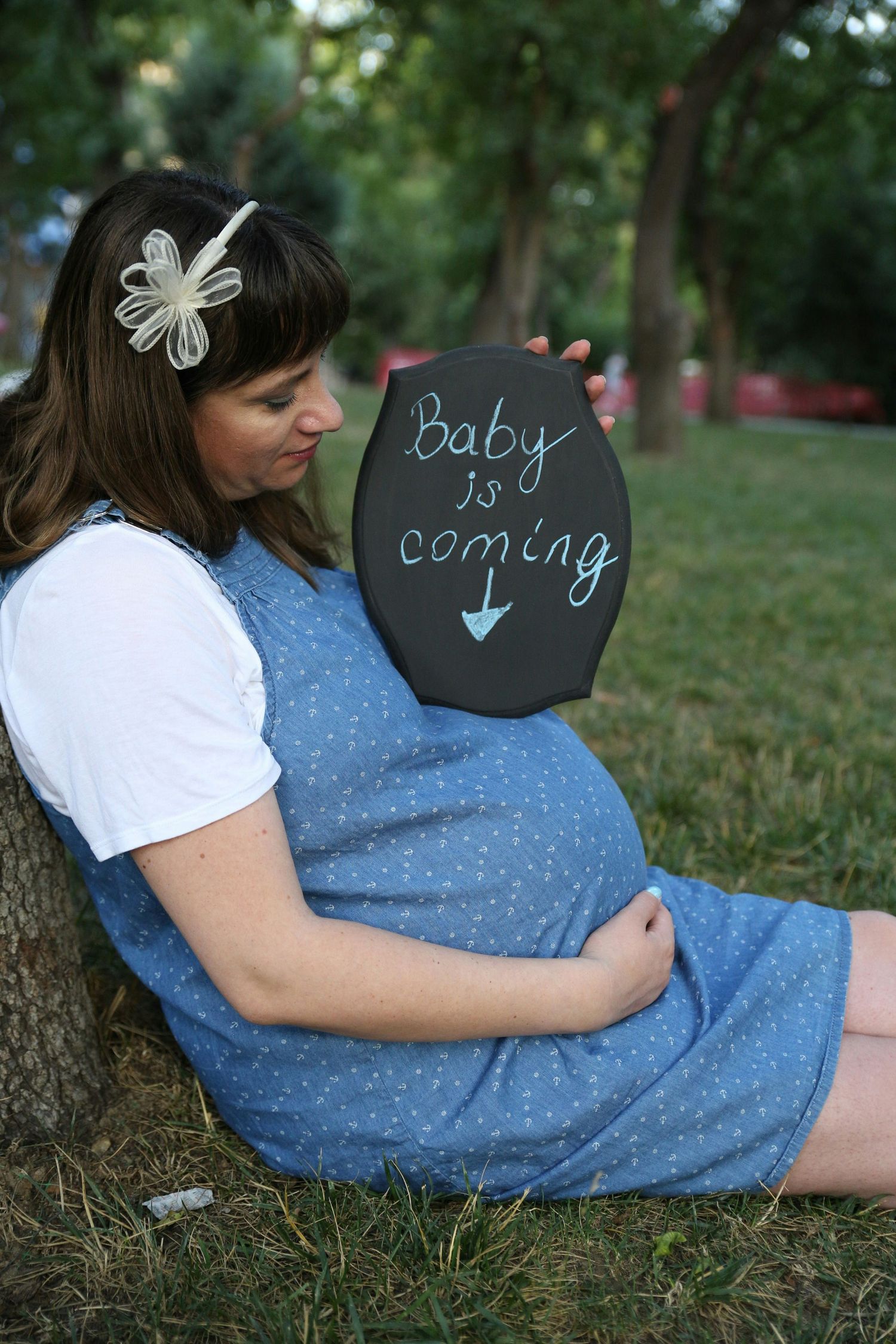 Pregnant woman leaning against tree holding baby is coming chalkboard sign.