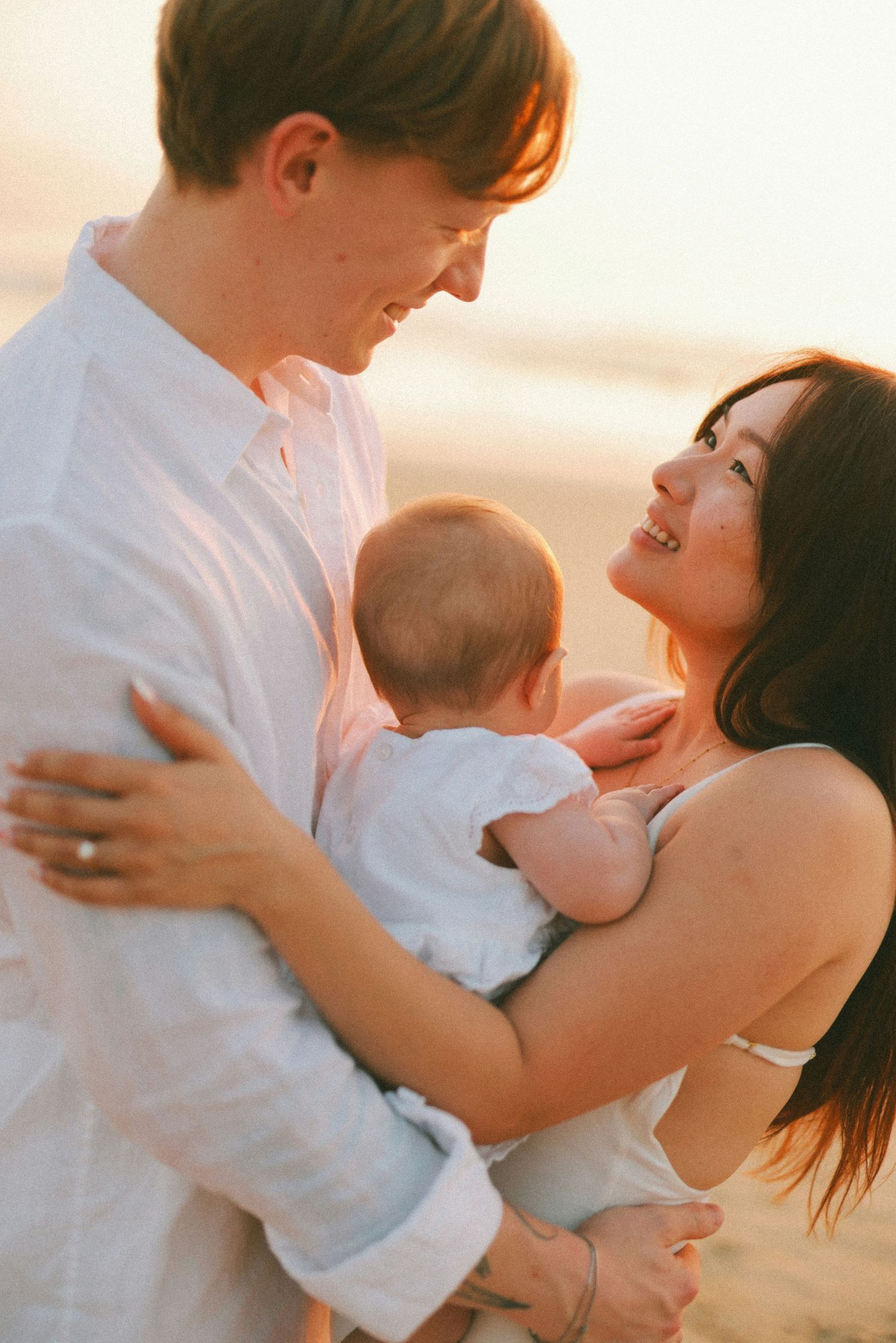 Happy couple smiling at each other while holding their newborn baby on a sunlit beach at golden hour.