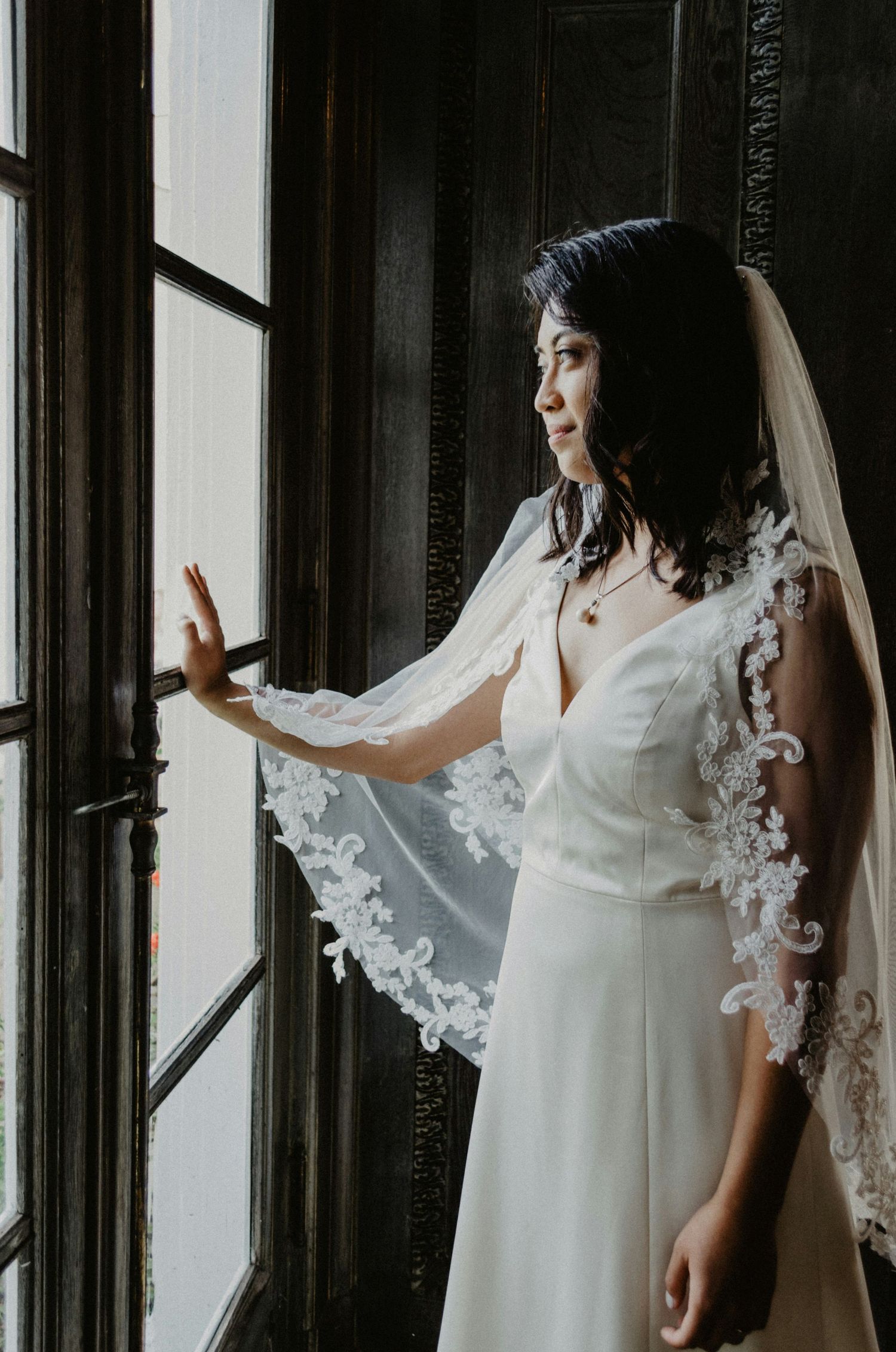 Beautiful bride in white gown with lace-trimmed veil gazing out a dark wooden window frame.