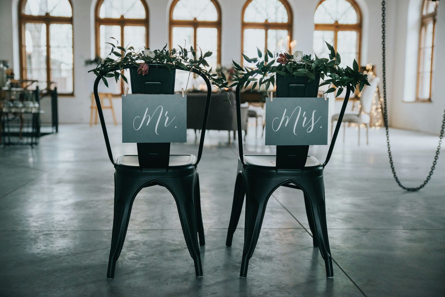 Black metal Mr. and Mrs. wedding chairs with greenery decorations in industrial venue with tall windows.