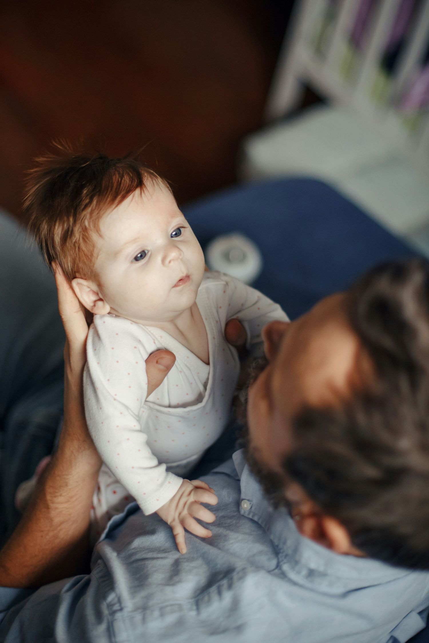 Curious red-haired baby in white onesie looking up attentively while being held by father on bed.