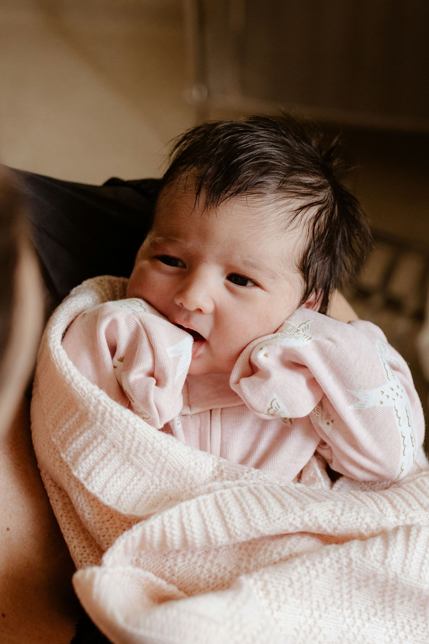 Newborn baby wrapped in pink blanket being held, with dark hair and alert eyes.