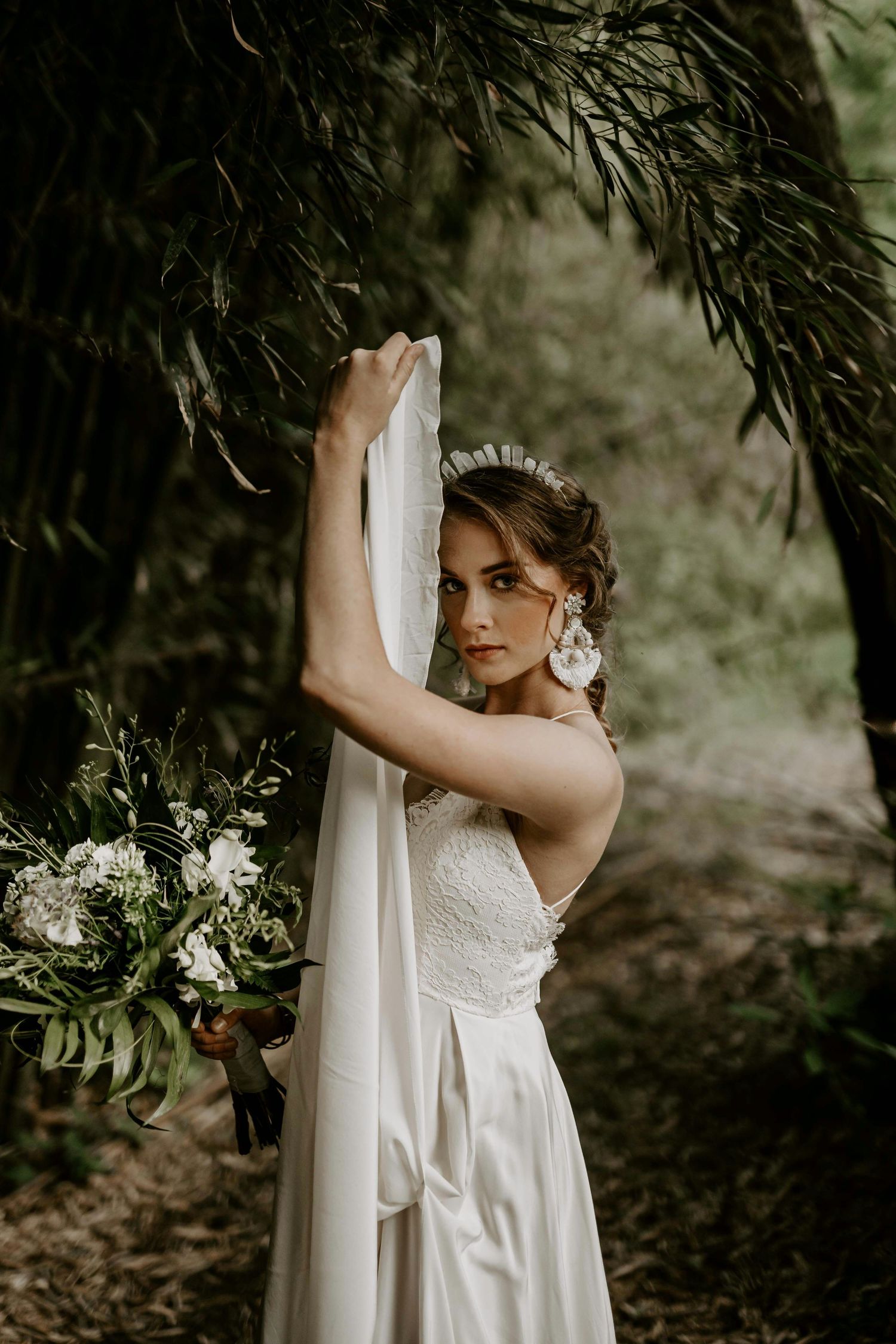 Bride in white satin gown holds veil and bouquet among green bamboo trees.