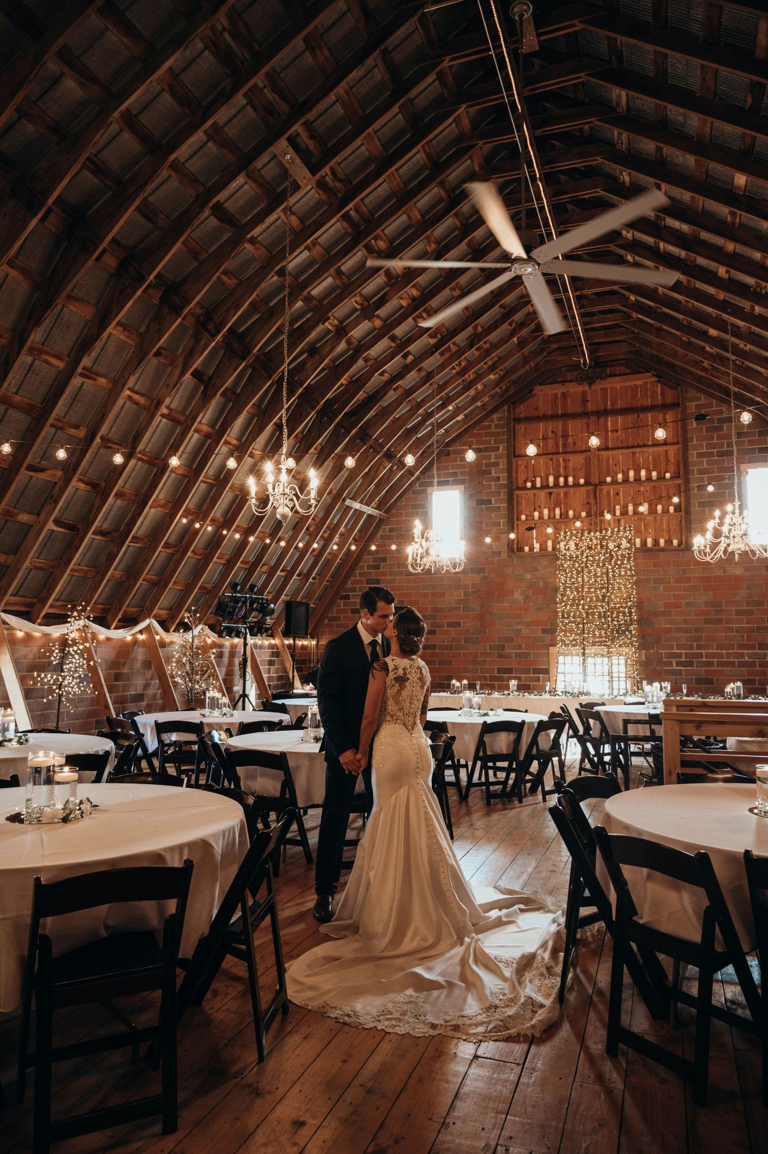 Bride and groom share an intimate moment inside a rustic barn wedding venue with chandeliers and string lights.