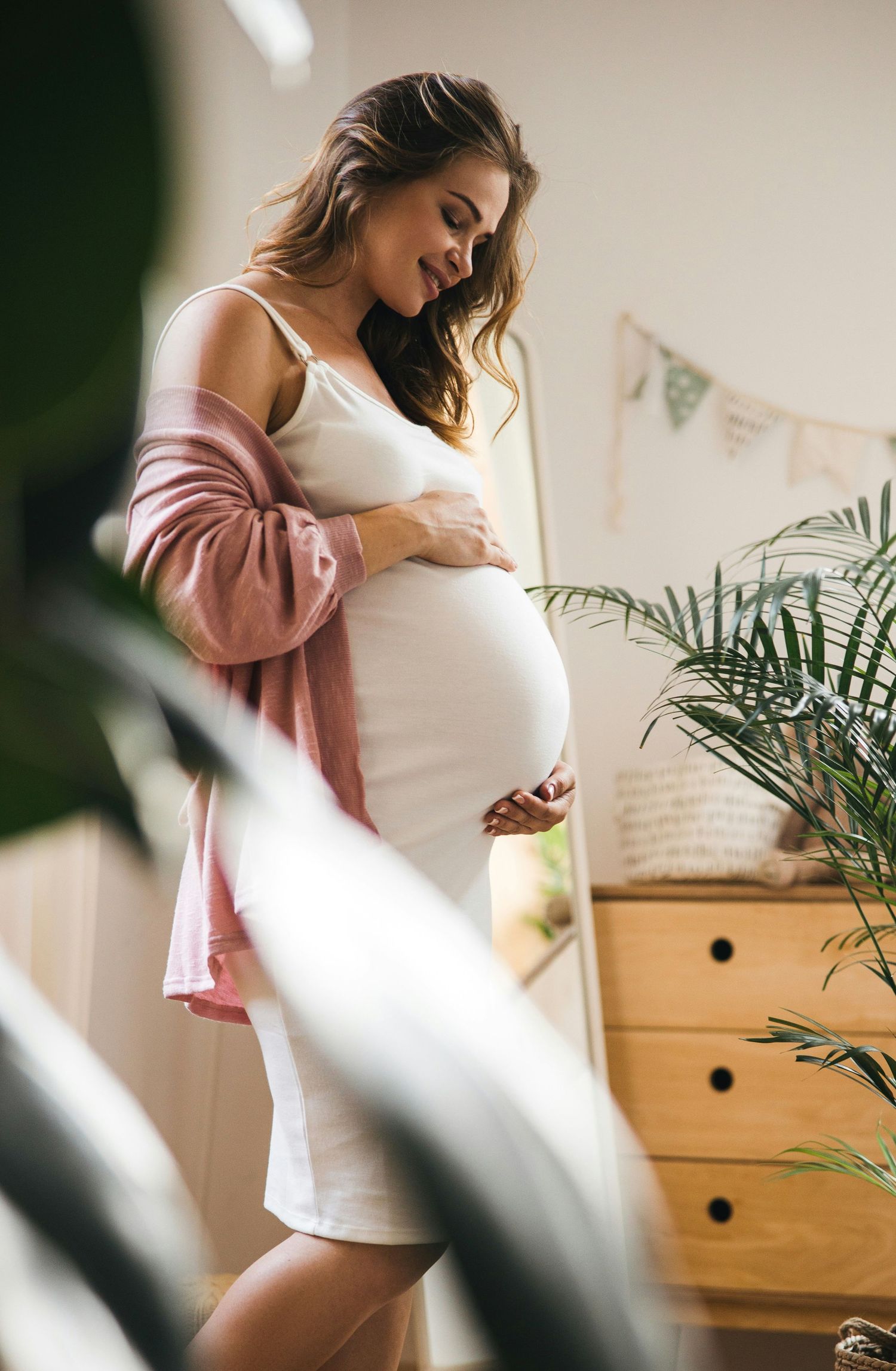 A pregnant woman in a white dress and pink cardigan gently cradling her baby bump in a cozy nursery.