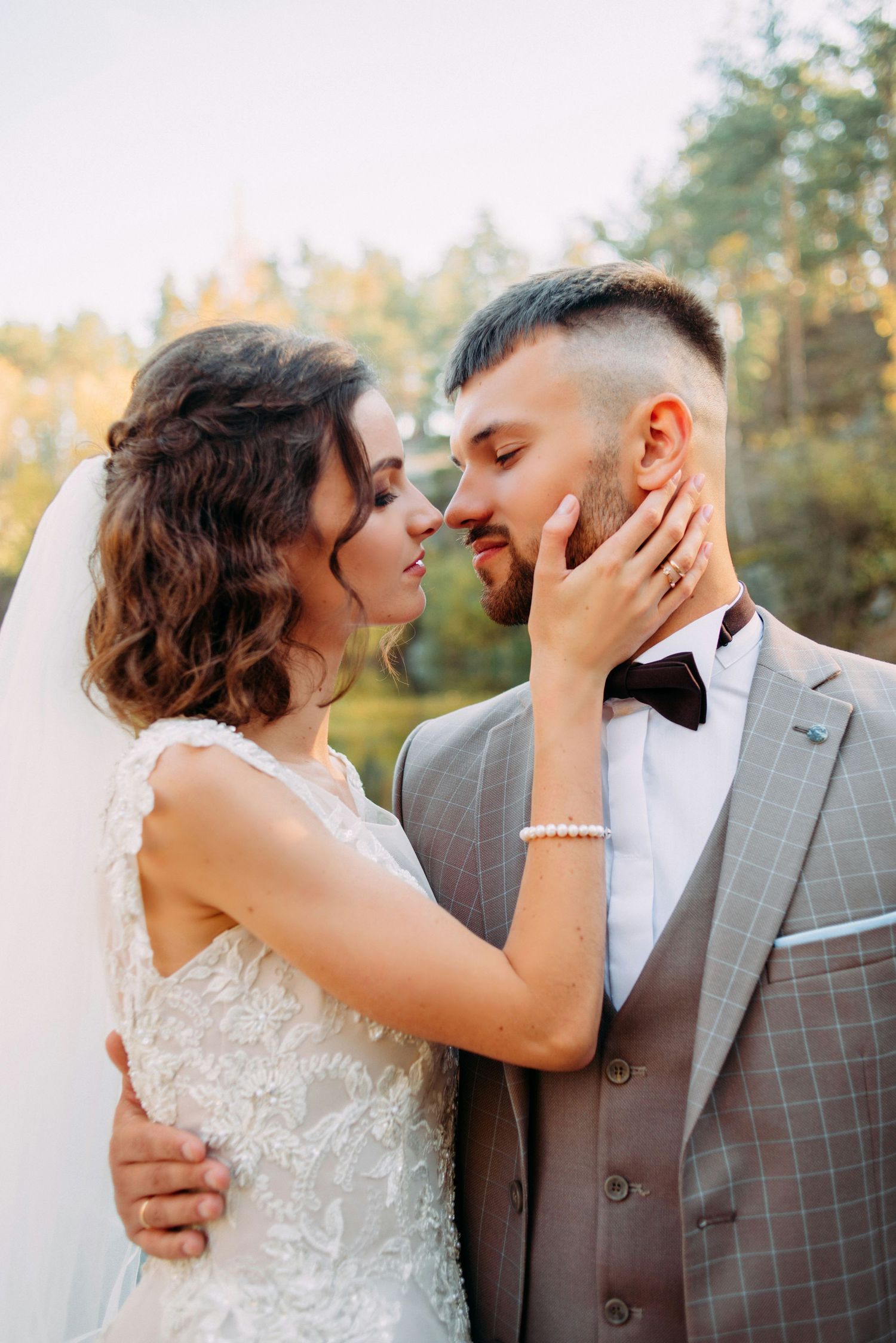 Bride in lace dress gently touching grooms face as they share an intimate moment outdoors in sunlight.