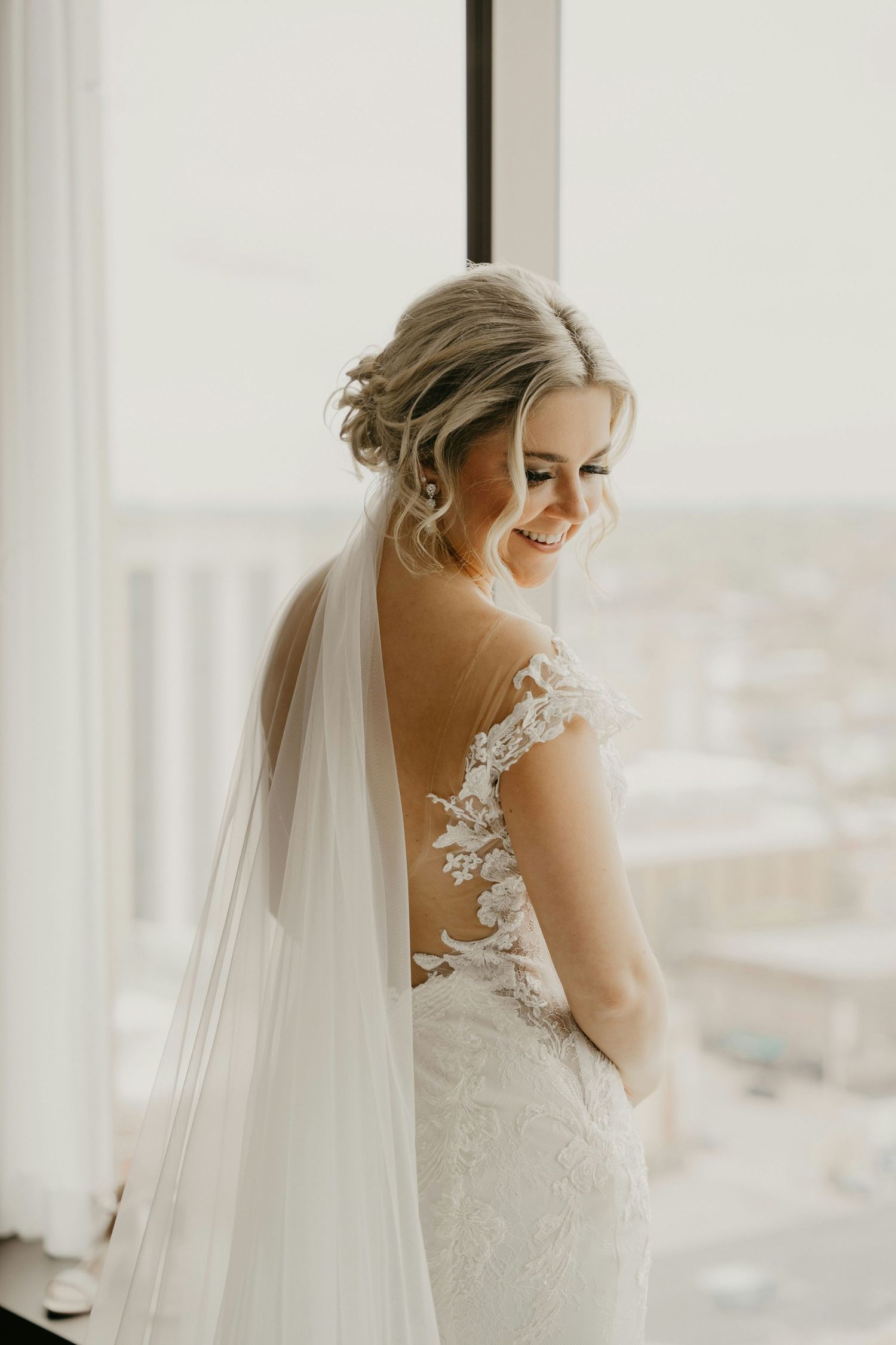 Smiling bride in lace backless wedding gown with veil standing by large window.