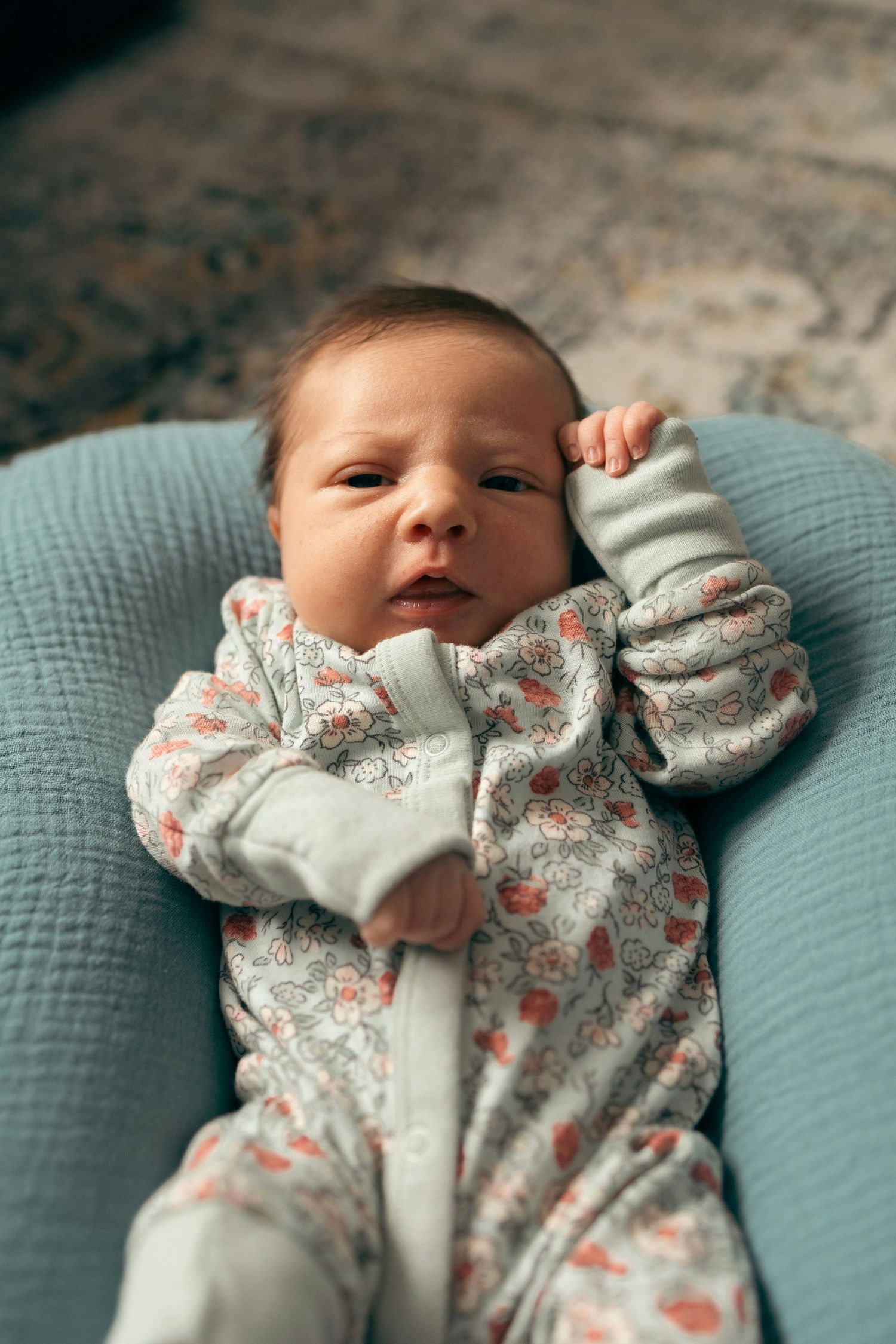 Alert newborn baby in a floral zip-up sleeper lying awake in a light blue infant lounger.