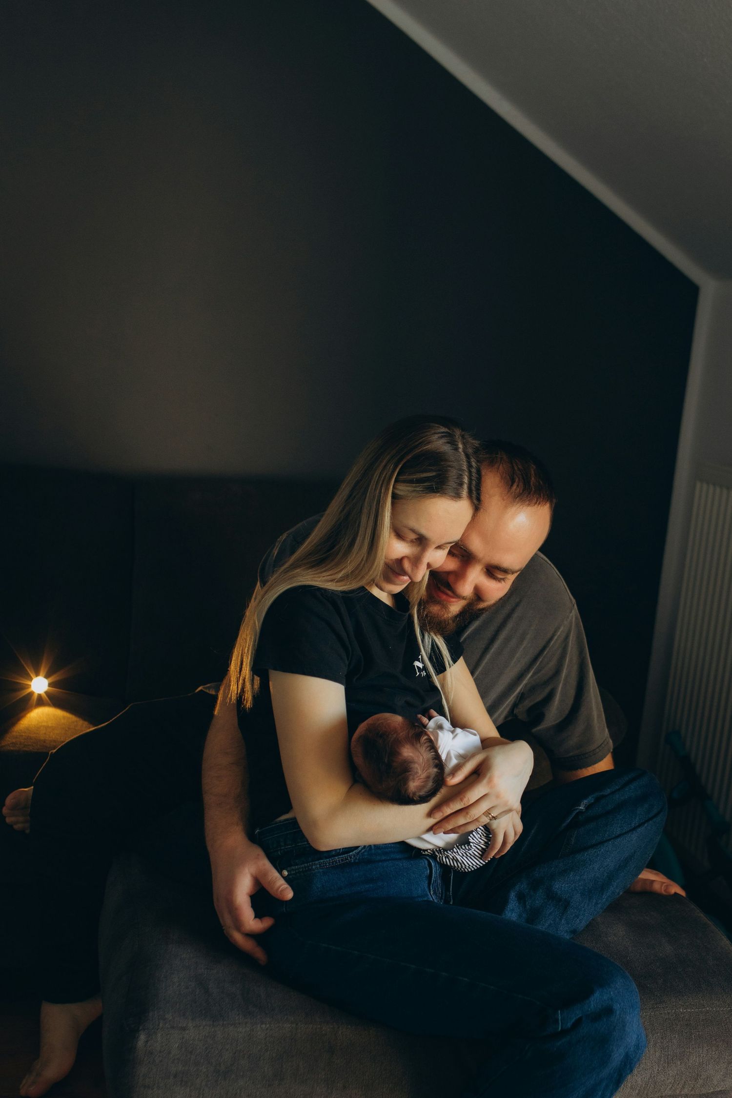 Couple sitting together in dimly lit room smiling at newborn baby they hold.