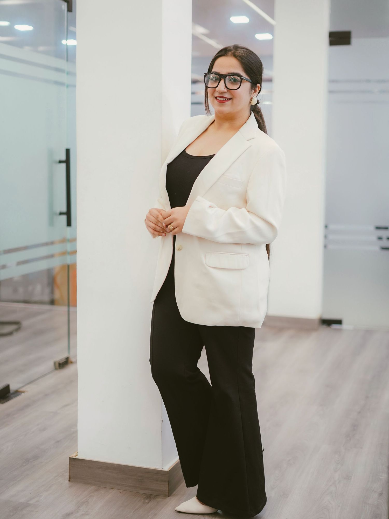 Woman in white blazer and black outfit leaning against white pillar in modern office setting.