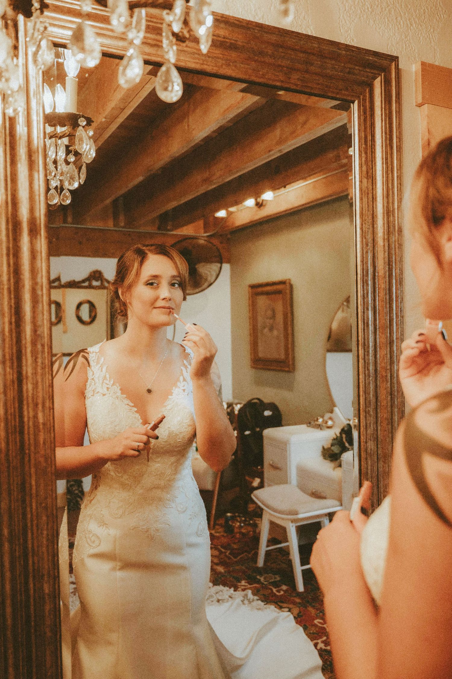 Bride in lace wedding gown applying makeup while gazing into ornate wooden mirror in elegant bridal suite.
