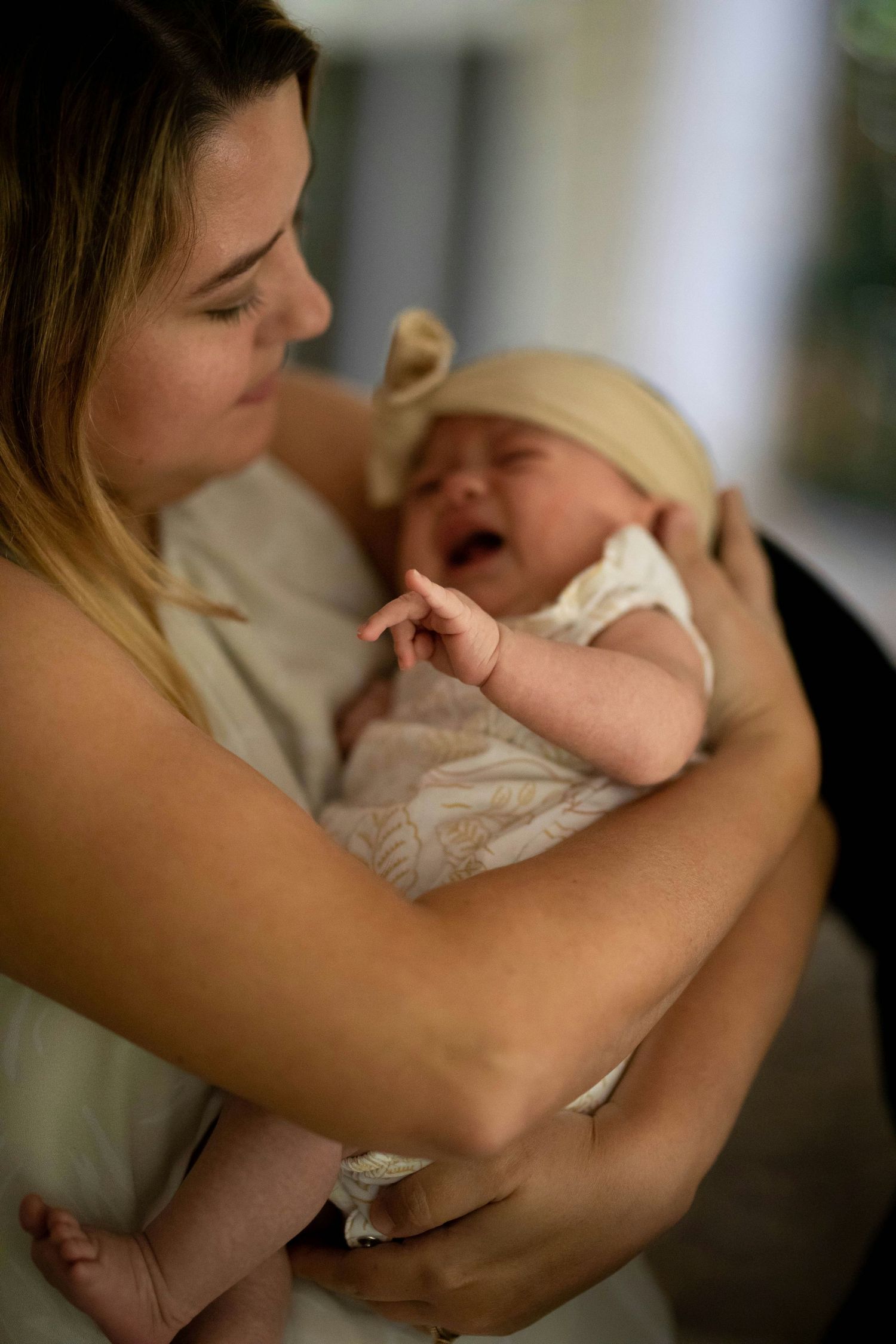 A young mother gently holds her crying newborn baby dressed in a cream outfit with a bow headband.
