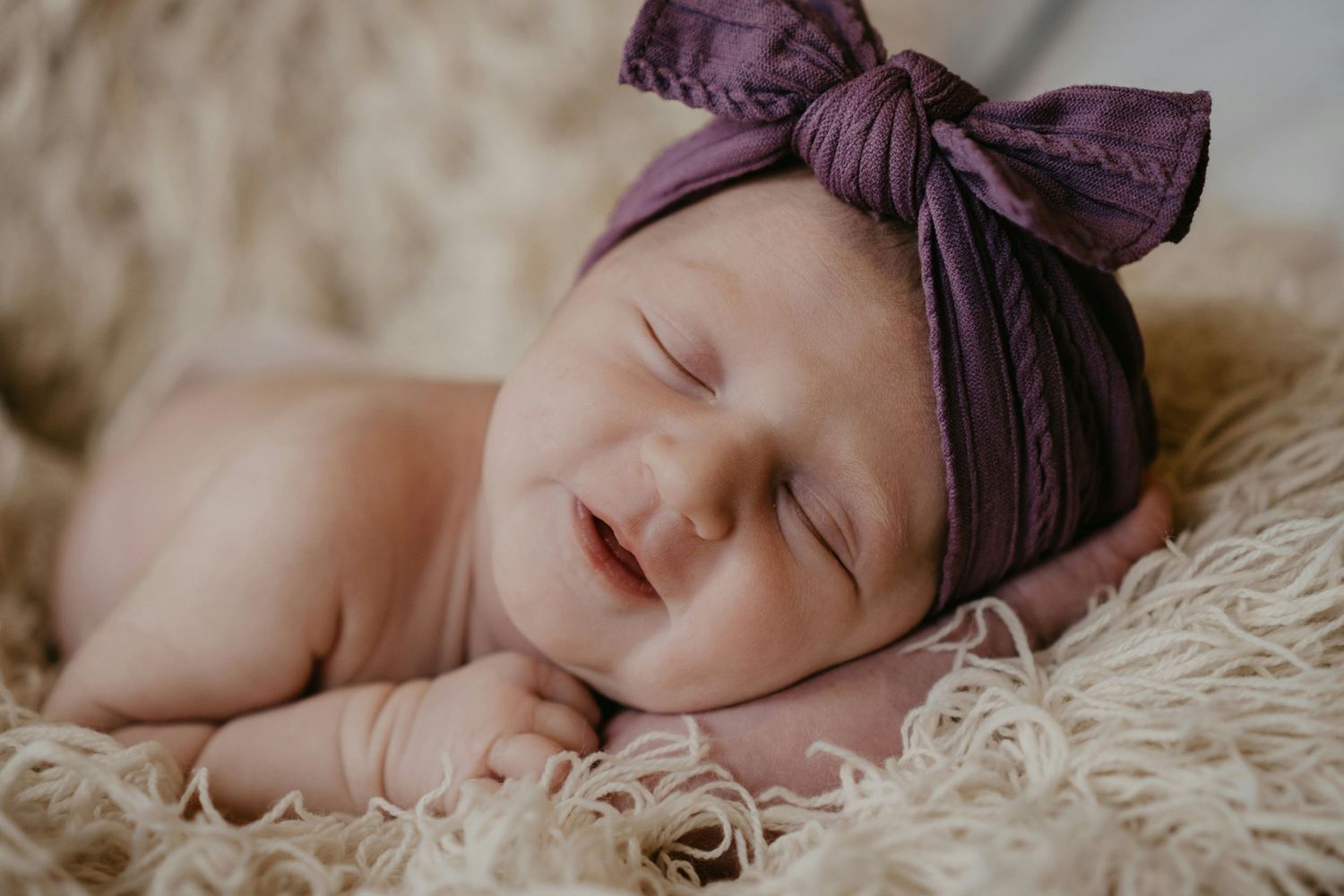 Smiling newborn baby wearing purple bow headband resting on fluffy beige textured surface.
