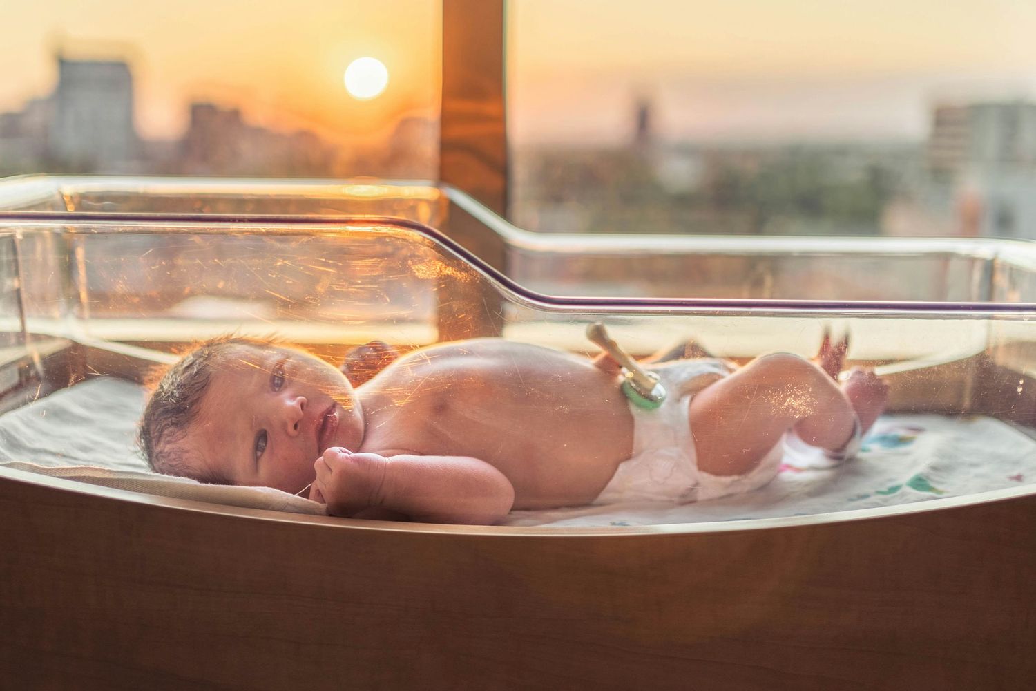 Newborn baby lying in hospital bassinet with golden sunset cityscape visible through window.