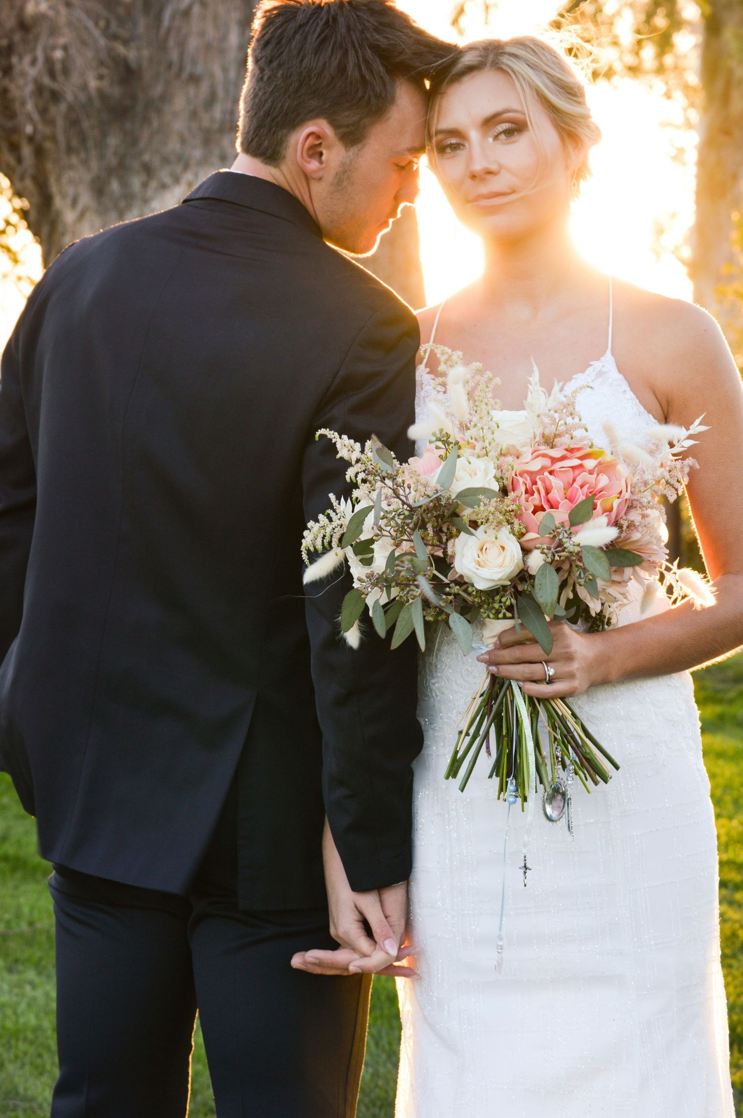 Bride holding pink and white bouquet leans into groom in black suit during golden hour outdoor wedding portrait.