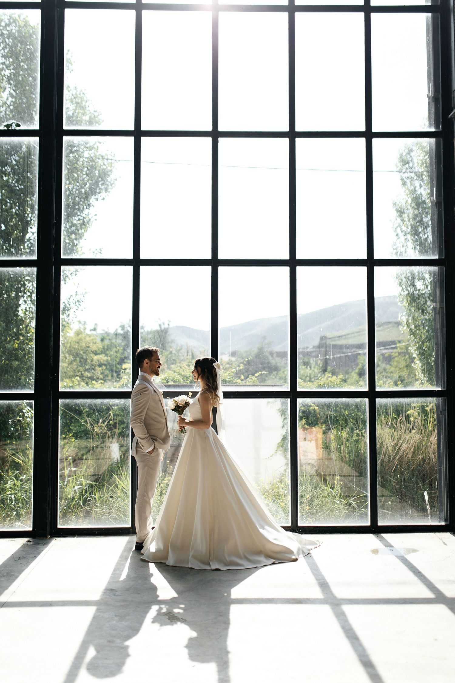 Bride and groom standing before large industrial windows with mountain view in sunlit wedding venue.
