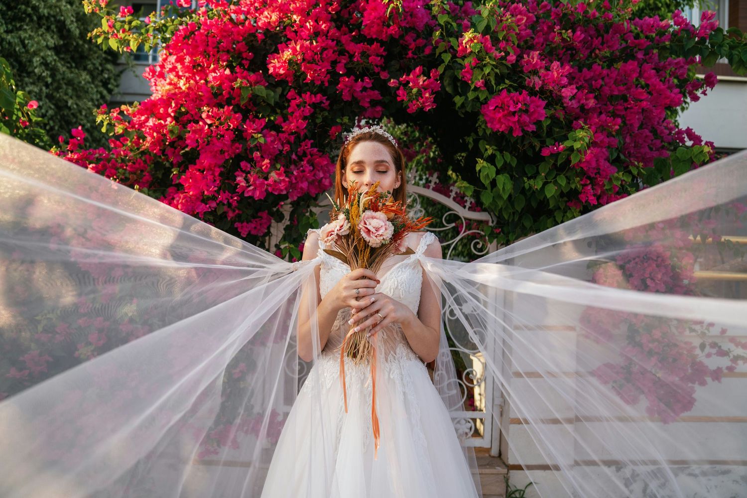 Bride in white gown holding bouquet with flowing veil against vibrant pink bougainvillea flowers.