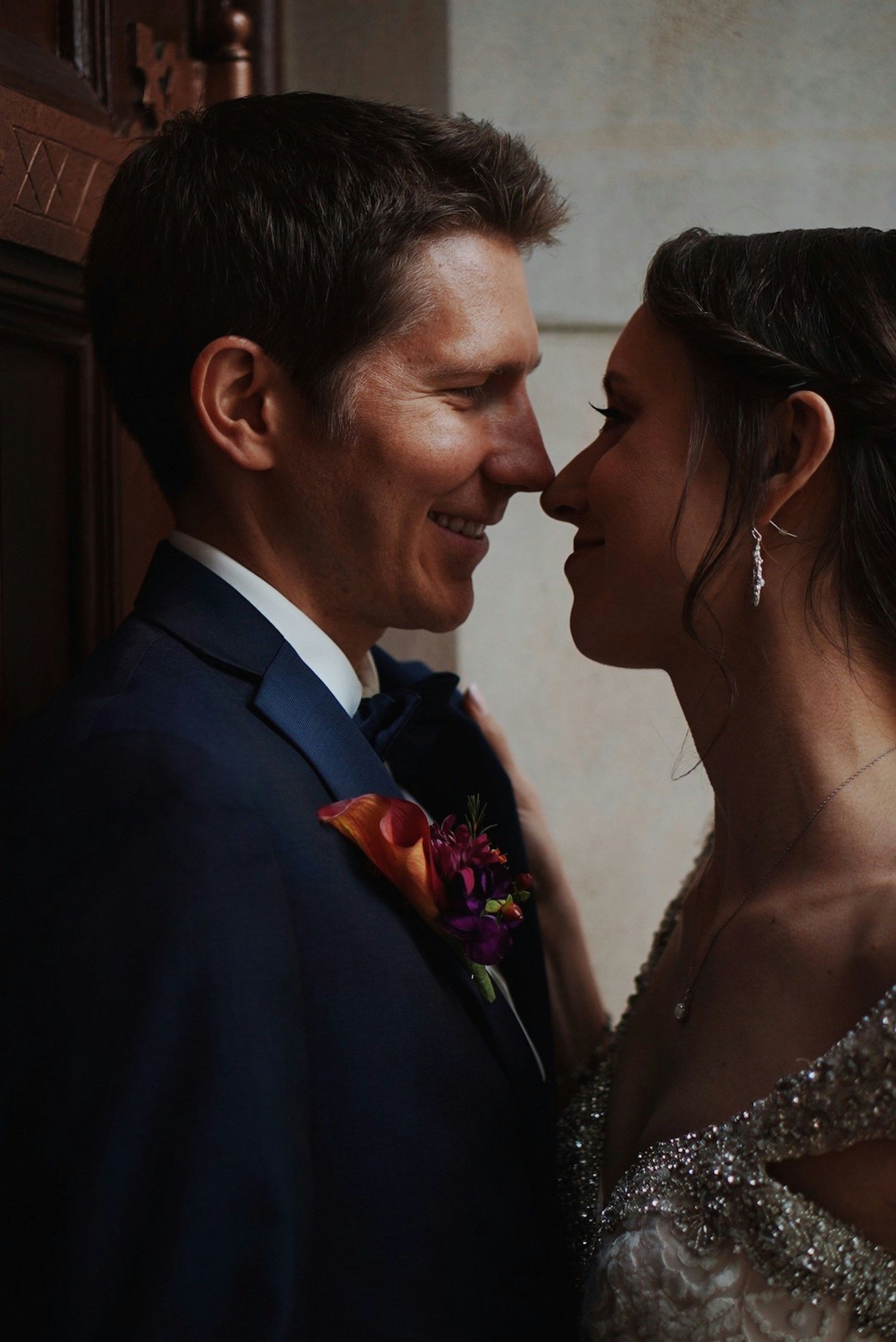 Couple shares an intimate nose-to-nose moment, groom in navy suit with floral boutonniere and bride in beaded gown.