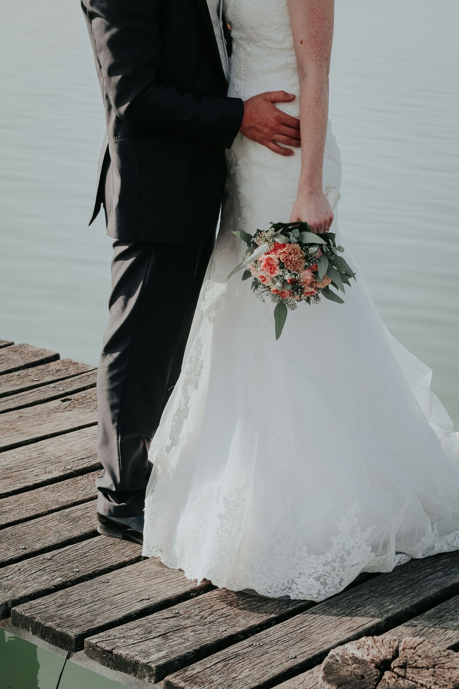 Bride and groom stand on a wooden dock by water, bride holding a pink and white wedding bouquet.