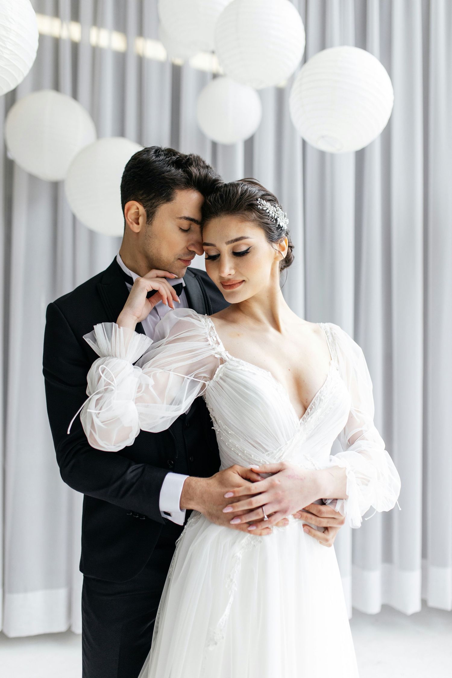 Romantic couple embracing in white decorated venue, bride in deep V-neck gown with puff sleeves.