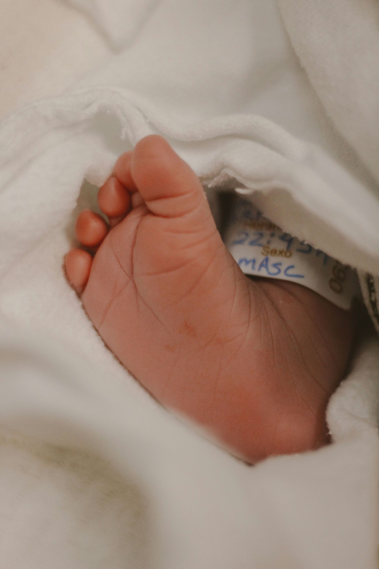 Newborn baby foot with hospital identification tag wrapped in white blanket.