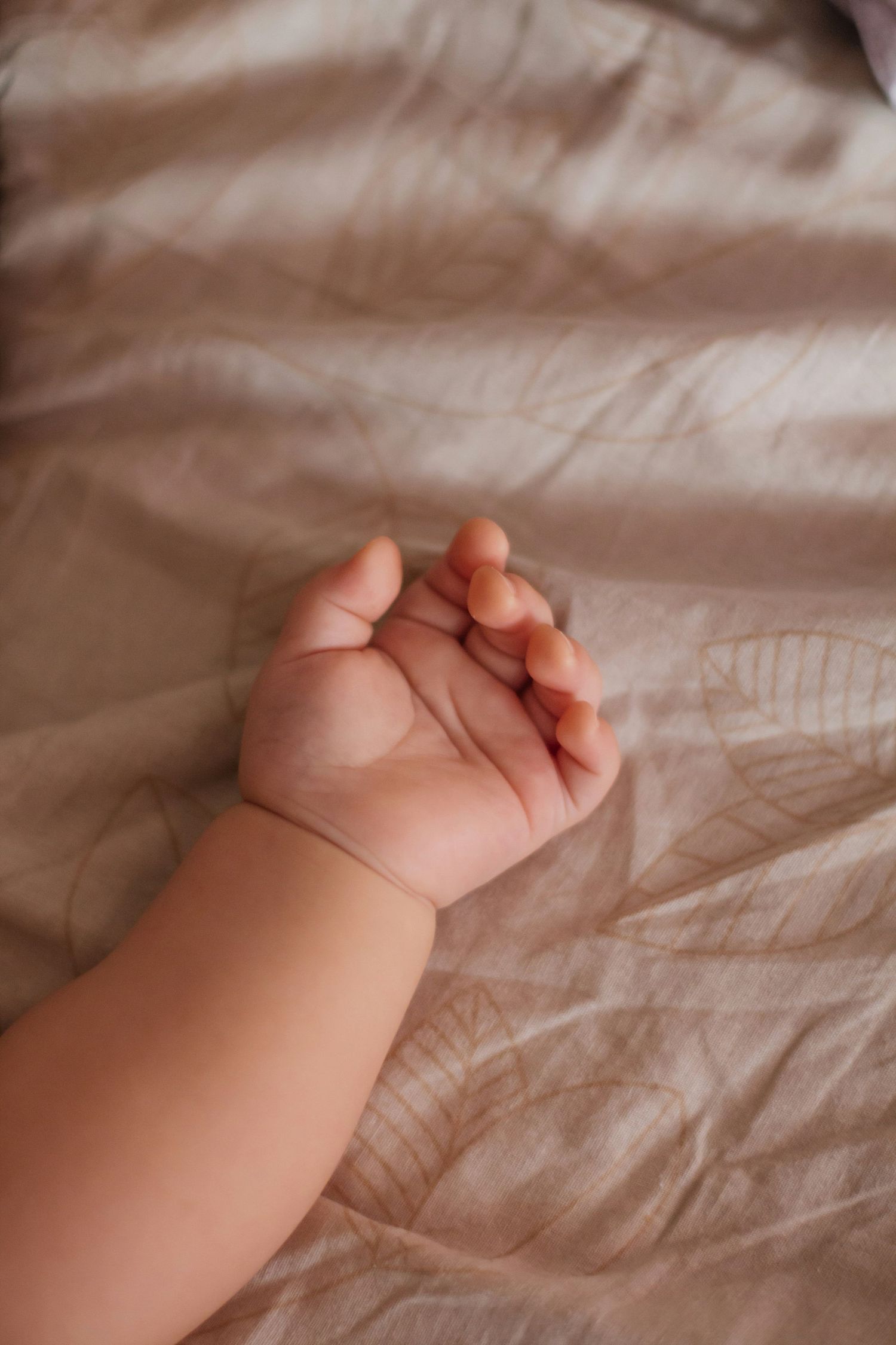 Close-up of a small baby hand resting gently on a beige leaf-patterned blanket.