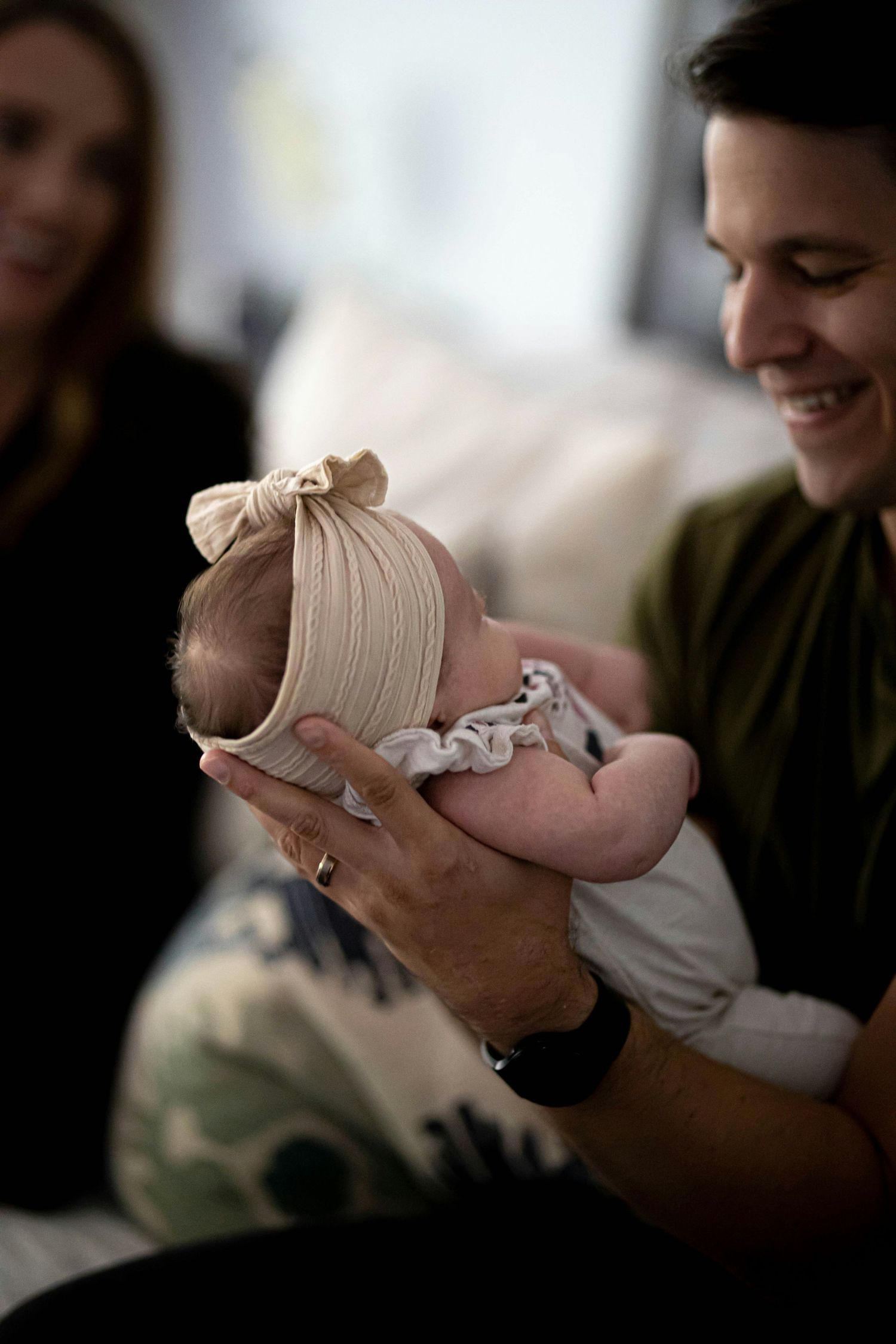 A smiling father carefully holds his tiny newborn baby wearing a beige bow headband in his hands.