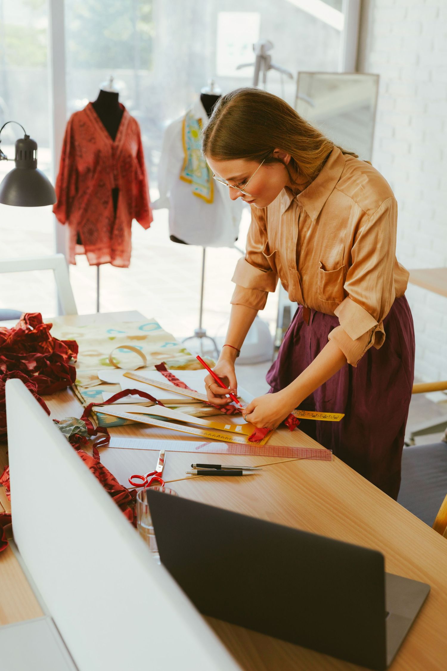 A female fashion designer cutting fabric with scissors on a worktable in a bright studio with mannequins in the background.