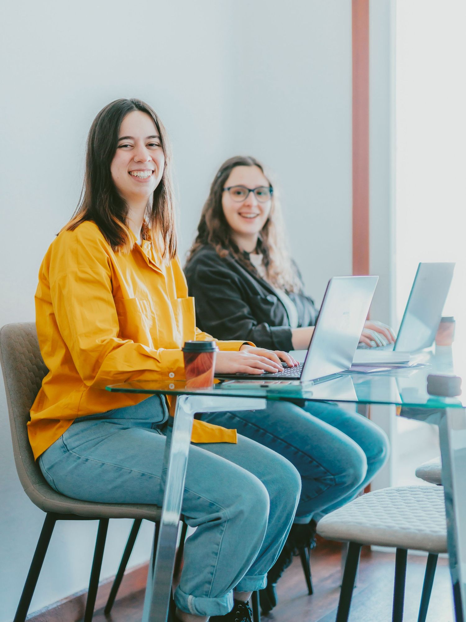 Two smiling women working on laptops at a glass desk in a bright modern office, one wearing a yellow shirt.