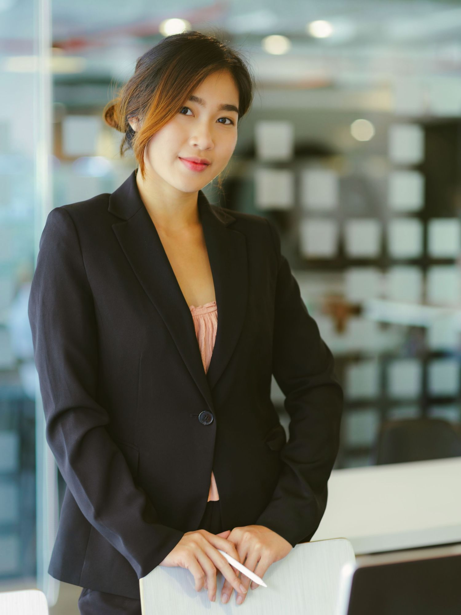 Professional Asian businesswoman in navy blazer holding pen, standing confidently in modern office environment.